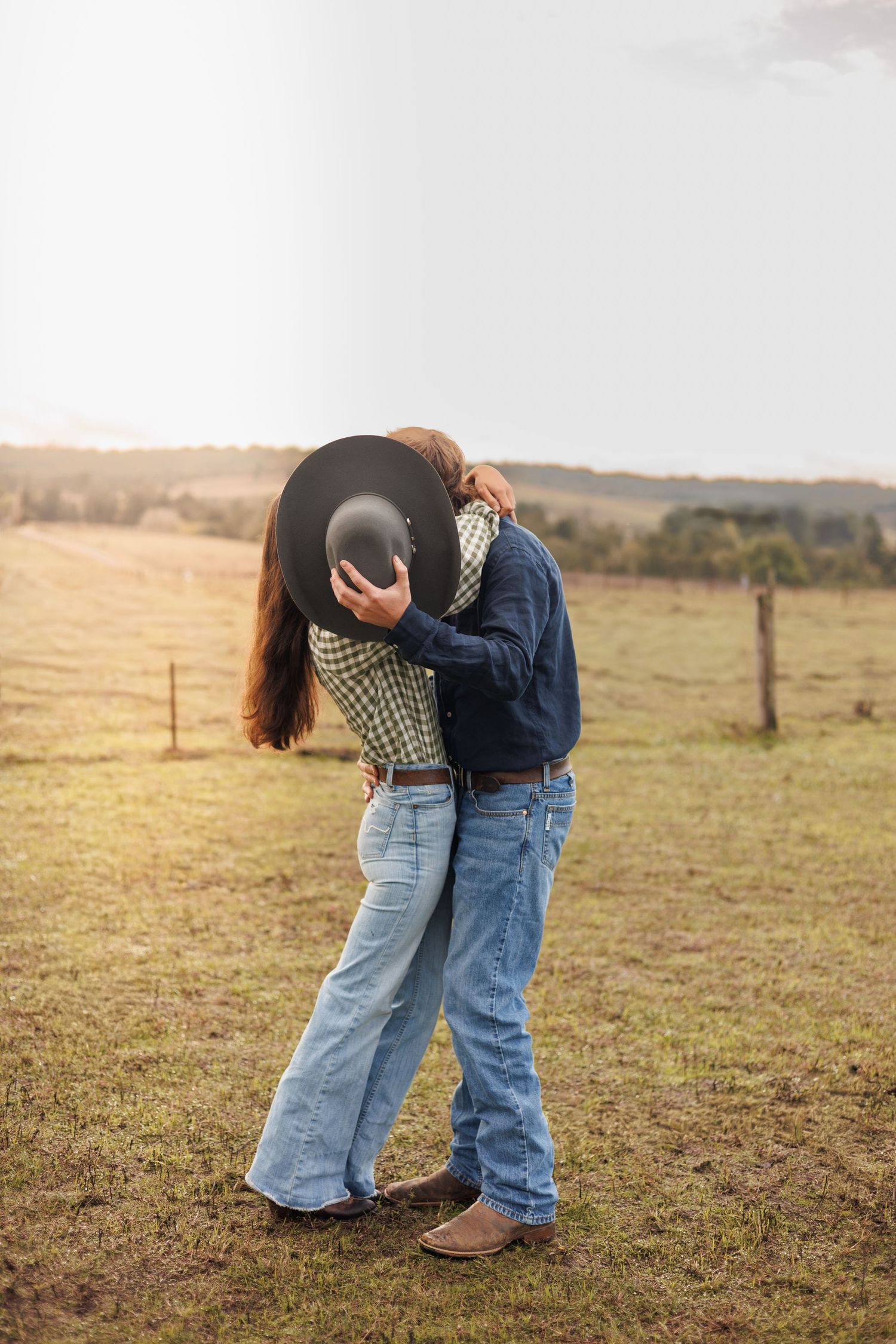 Couple kissing behind a black cowboy hat in a green field, wearing jeans and western attire at sunset.