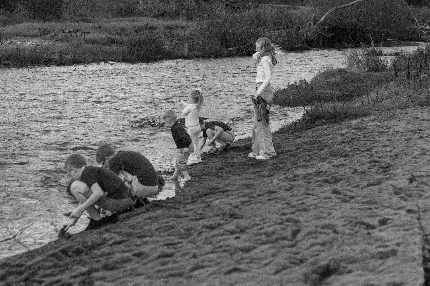 Children playing on a sandy riverbank in black and white photo, exploring nature by the water's edge.