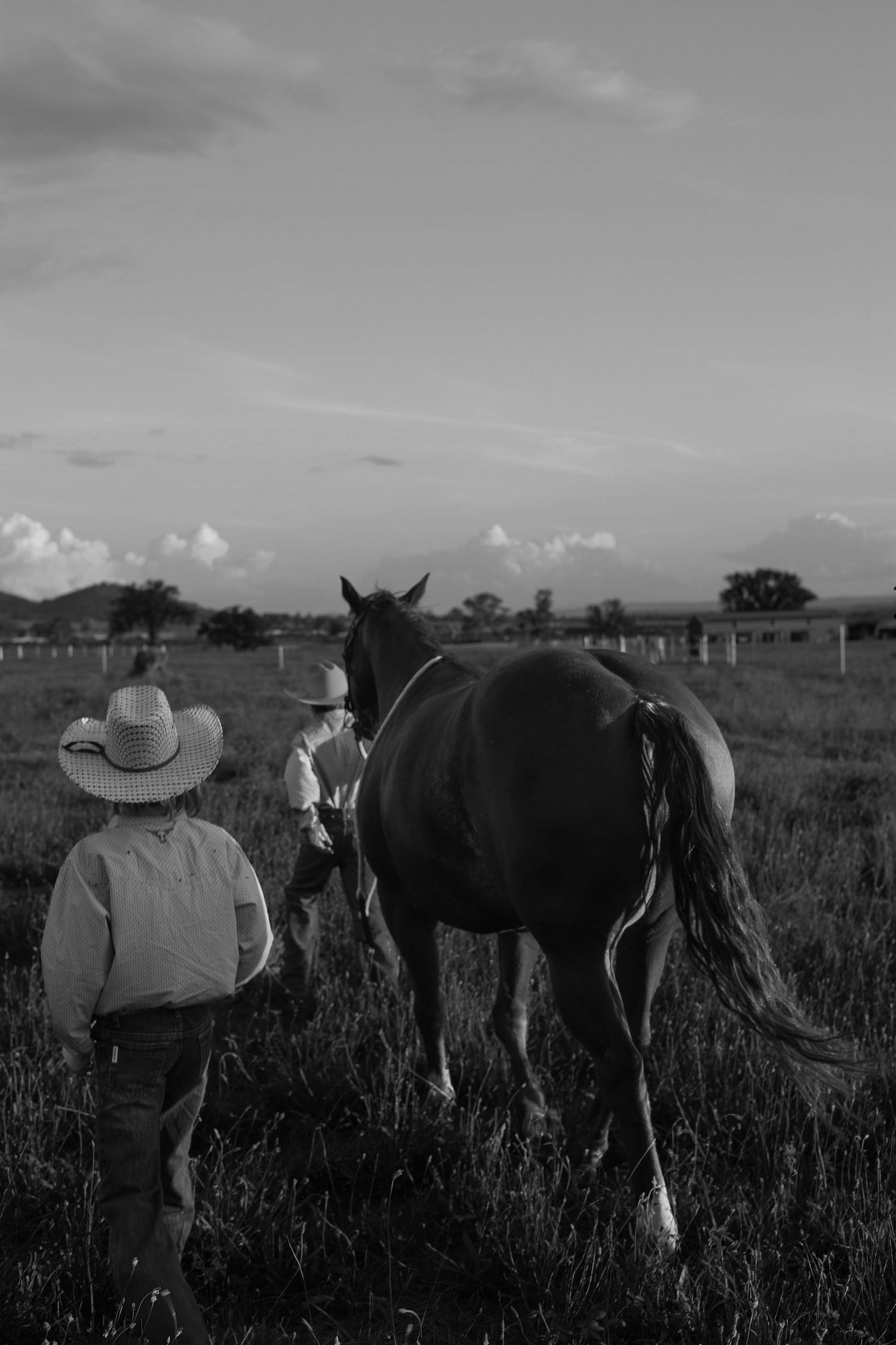 Black and white photo of two children in cowboy hats leading a horse across a grassy ranch field at dusk.