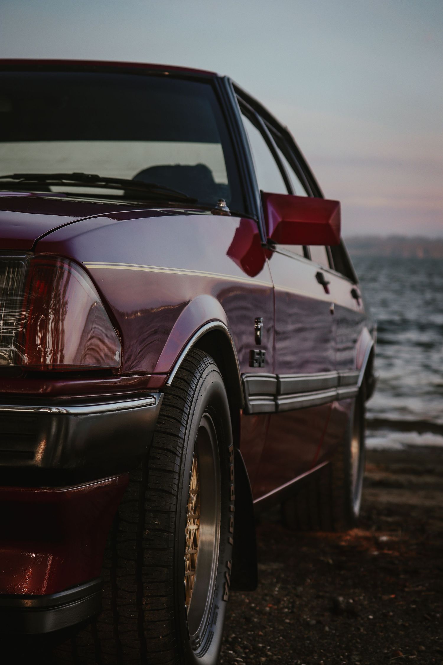 Low angle detail shots of a burgundy vintage car's wheels and body panels against a moody seaside backdrop at sunset.