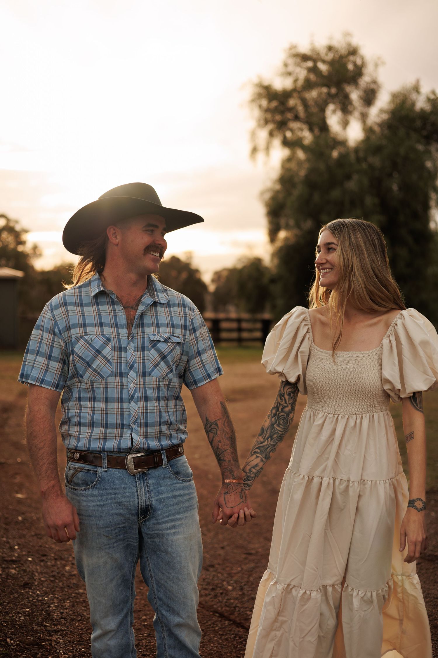A couple in western attire holds hands at sunset on a farm, man in cowboy hat and plaid shirt, woman in cream dress.