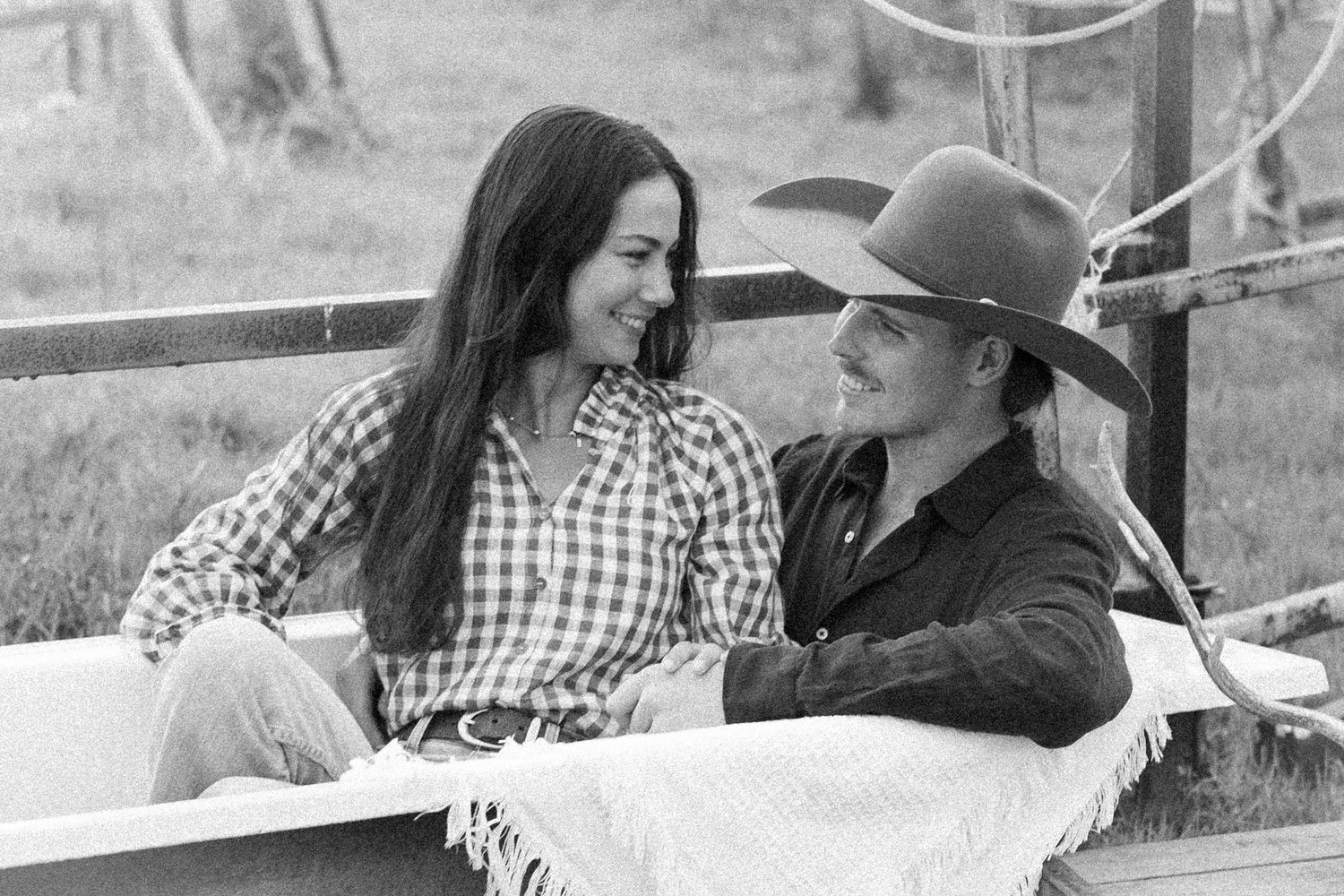 Black and white photo of a young couple in western wear smiling at each other outdoors on a ranch.
