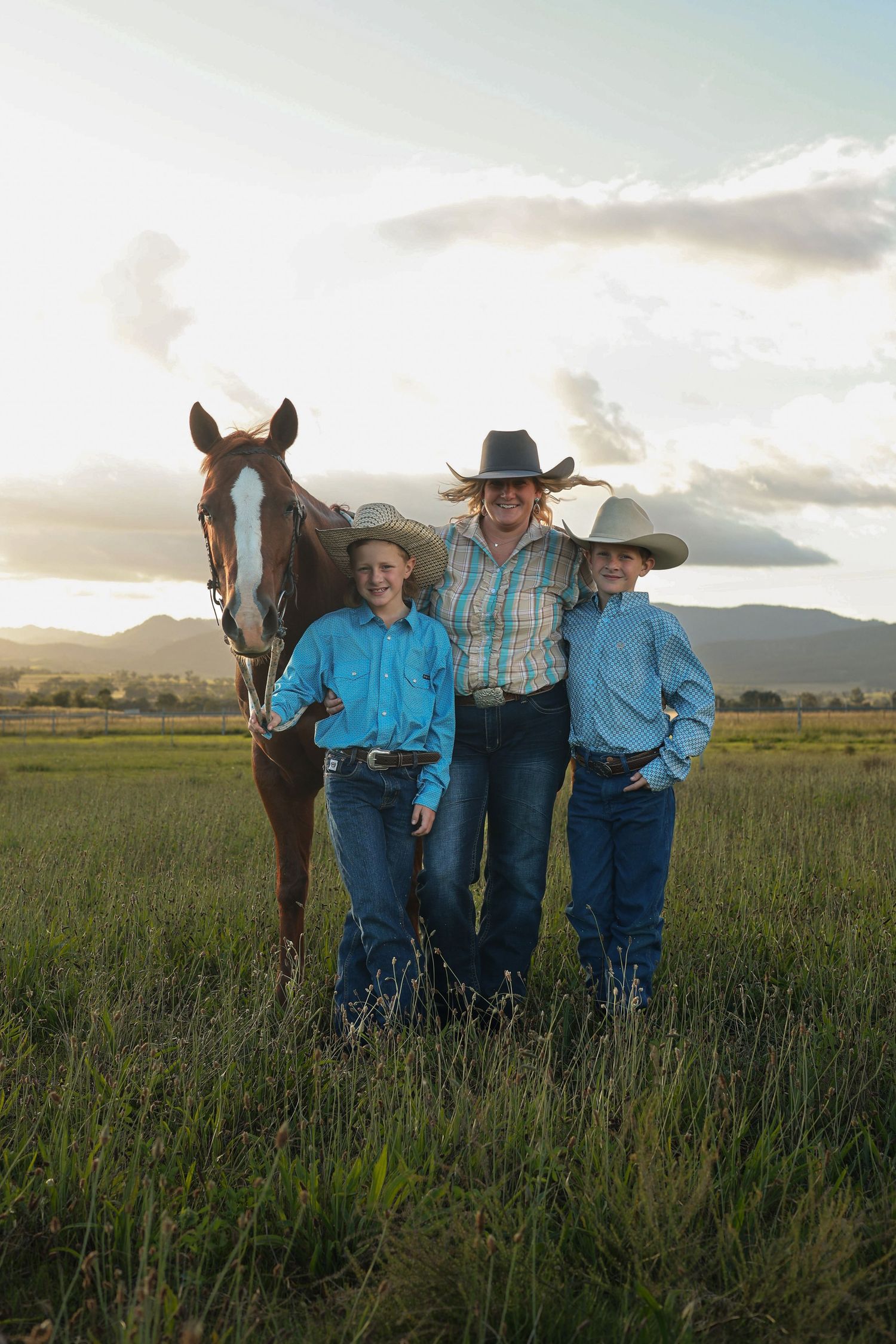 Three cowboys in blue shirts and hats pose with a brown horse in a green field at sunset.