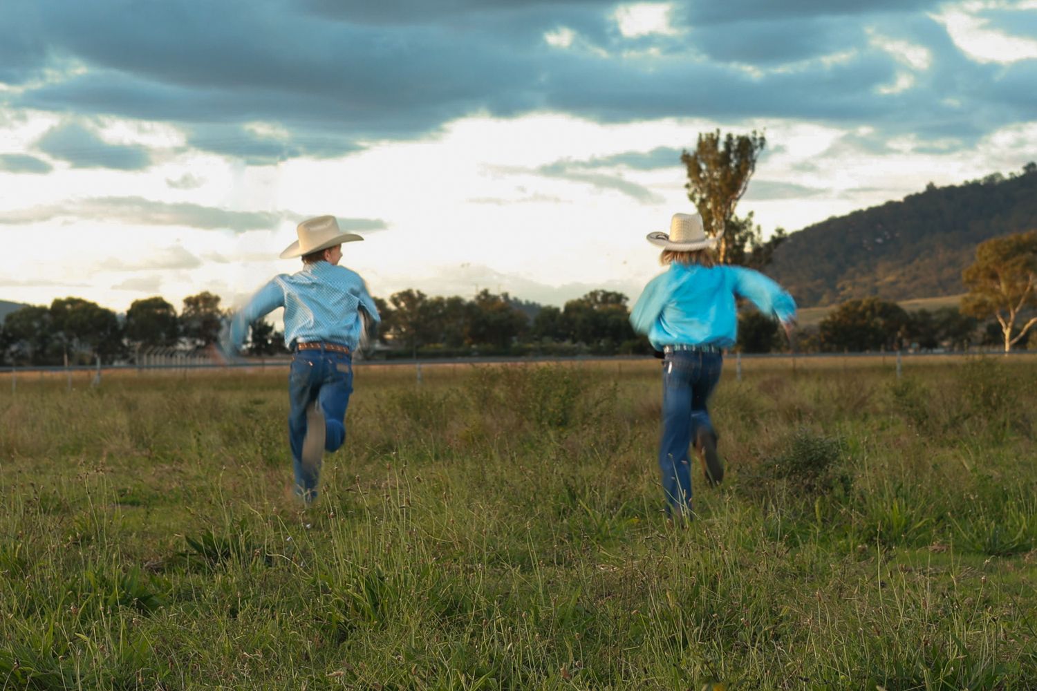 Two children in cowboy hats and blue shirts run through a green field under a dramatic cloudy sky.