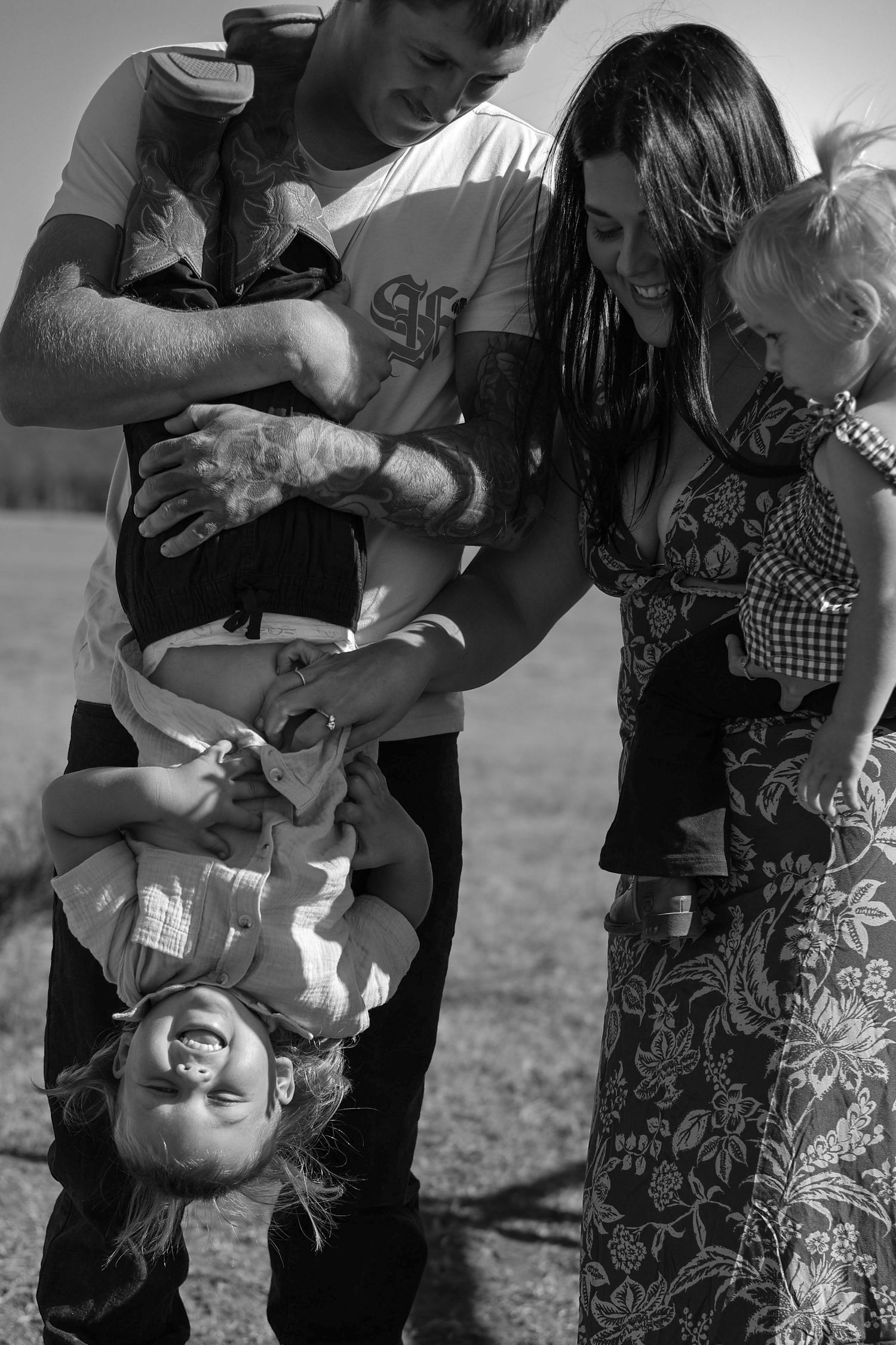 Black and white photo of a happy family, man holding child upside down while woman holds baby outdoors.