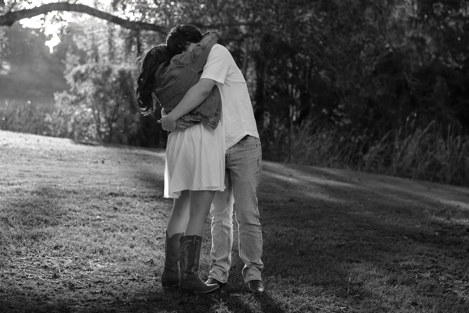 Black and white photo of a couple embracing tenderly on a grassy path surrounded by trees.