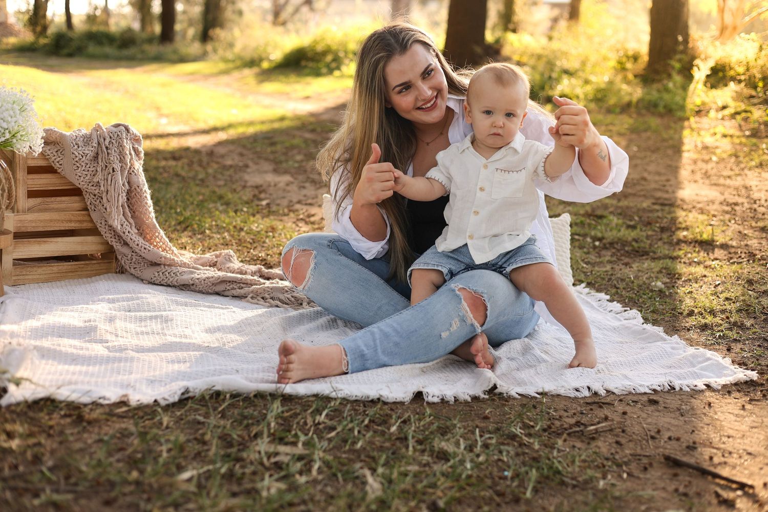 Young mother sitting on white blanket outdoors holding baby boy, both smiling and pointing in sunny park setting.