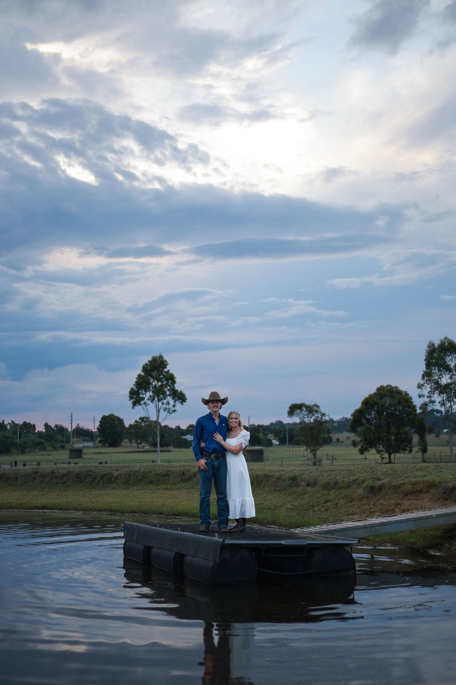 A couple stands together on a small dock overlooking calm waters at dusk with eucalyptus trees in the background.