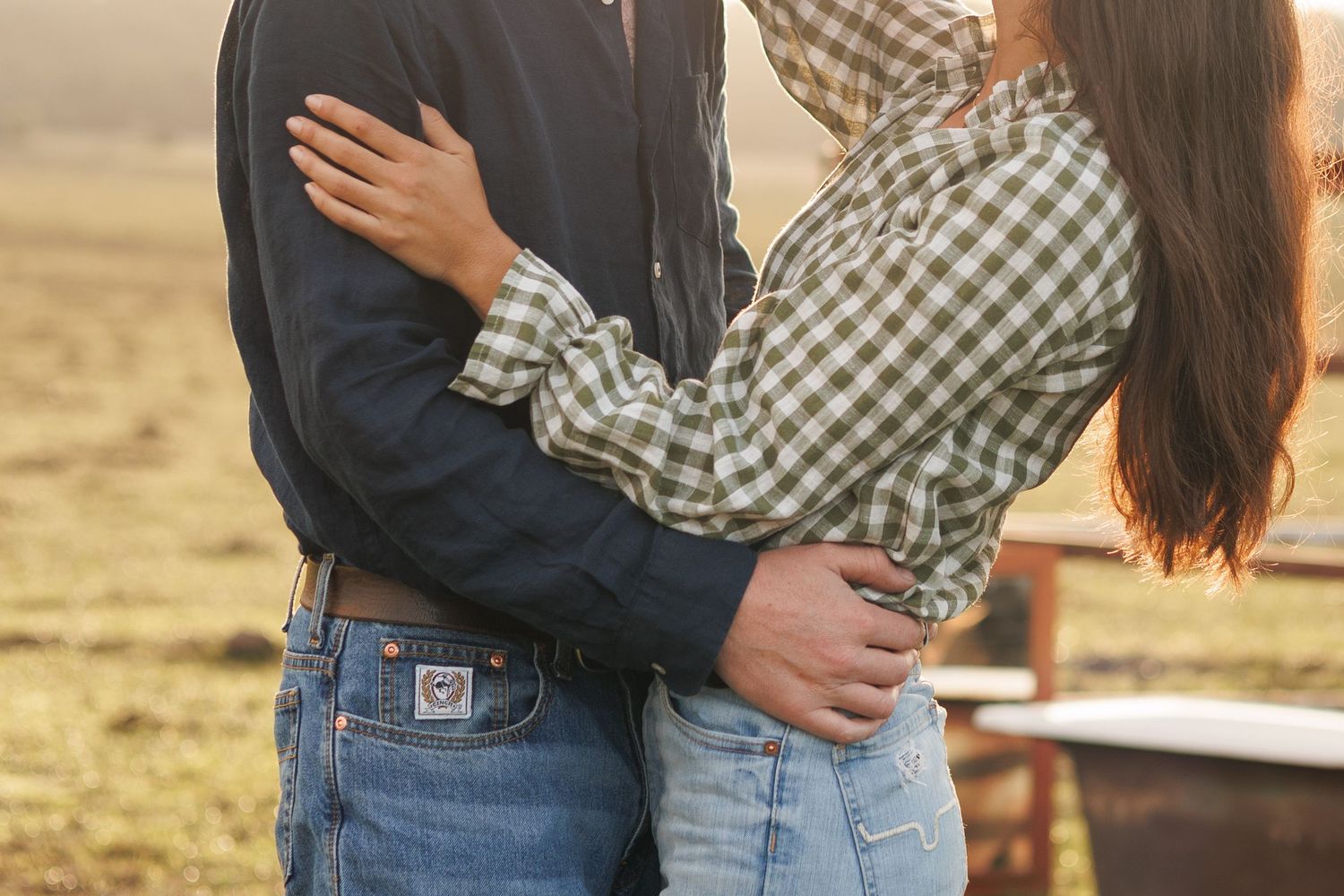 Couple embracing in a golden field at sunset, wearing jeans and plaid shirts in a romantic outdoor setting.