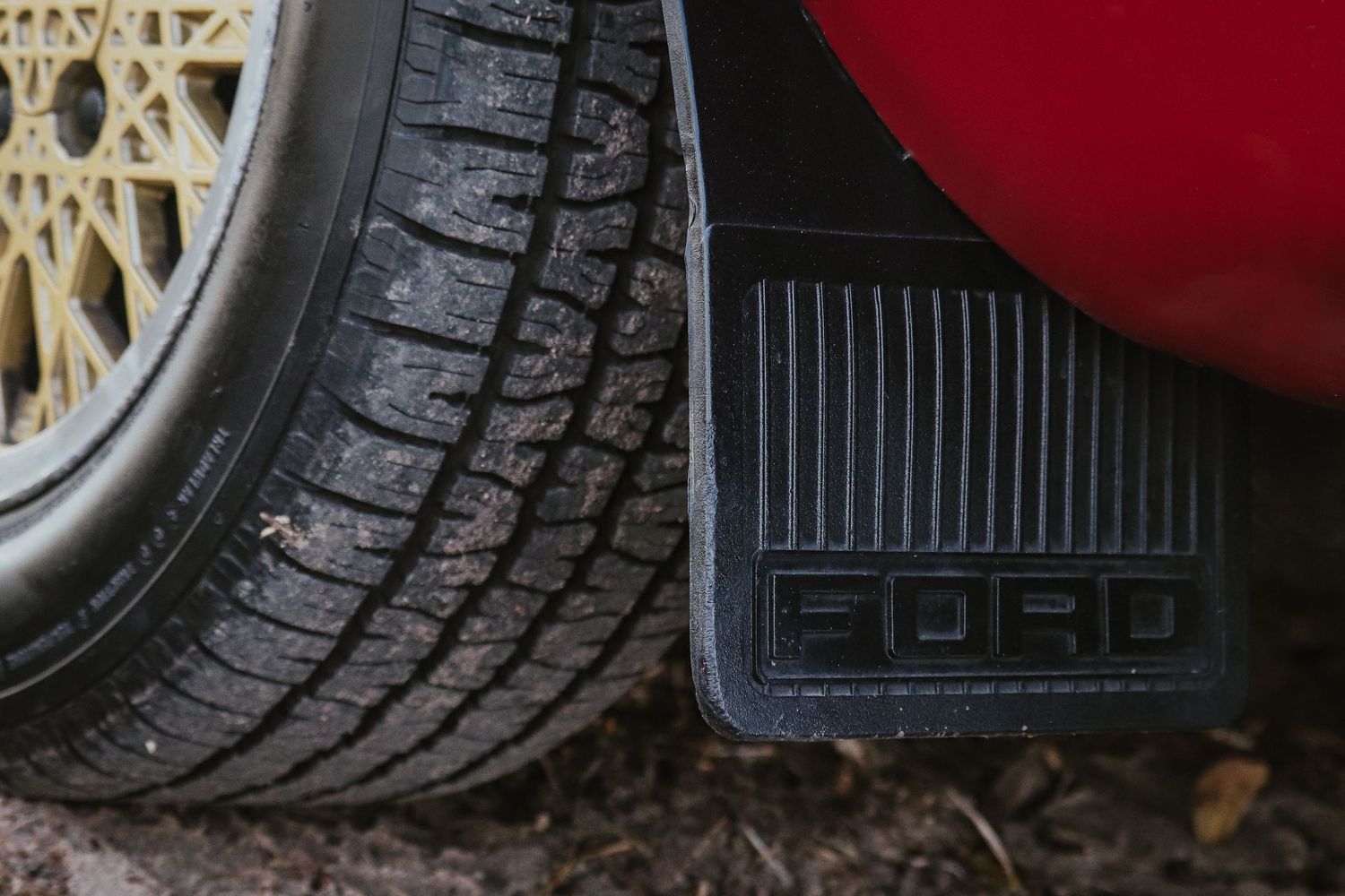 Close-up view of a car tire tread pattern next to a black Ford rubber mud flap on gravel.