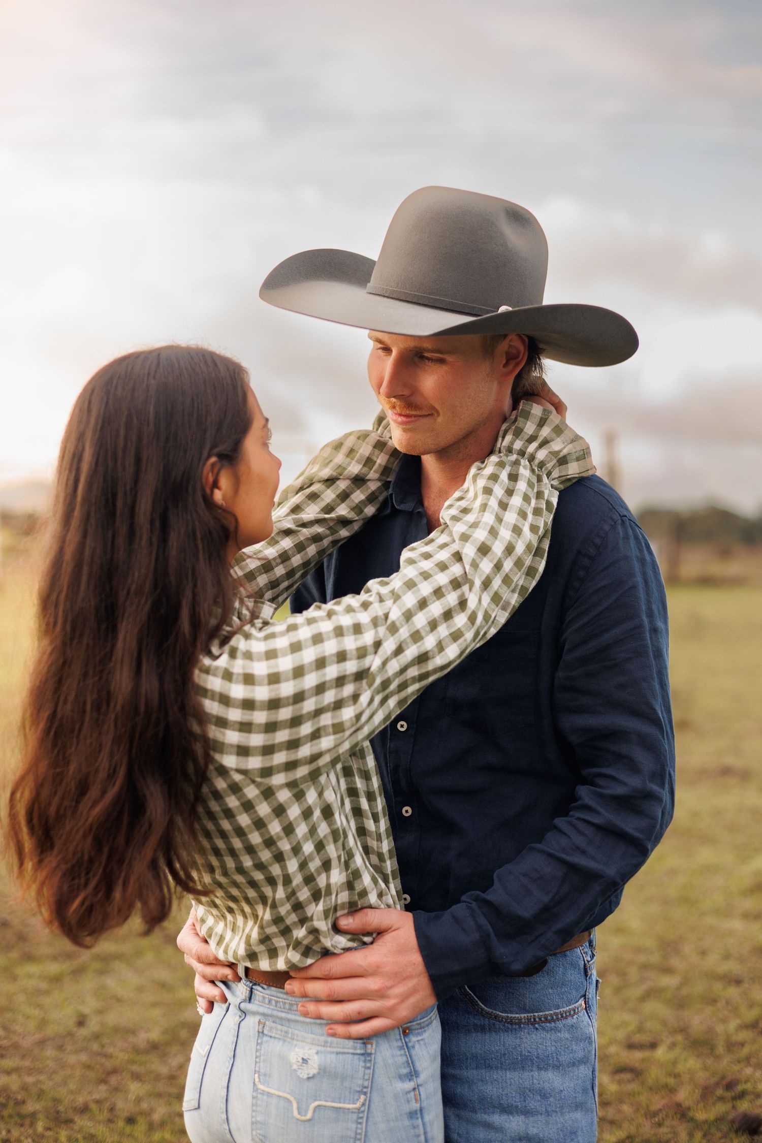 A couple embraces in a field, the man wearing a cowboy hat and blue shirt, the woman in a plaid top.