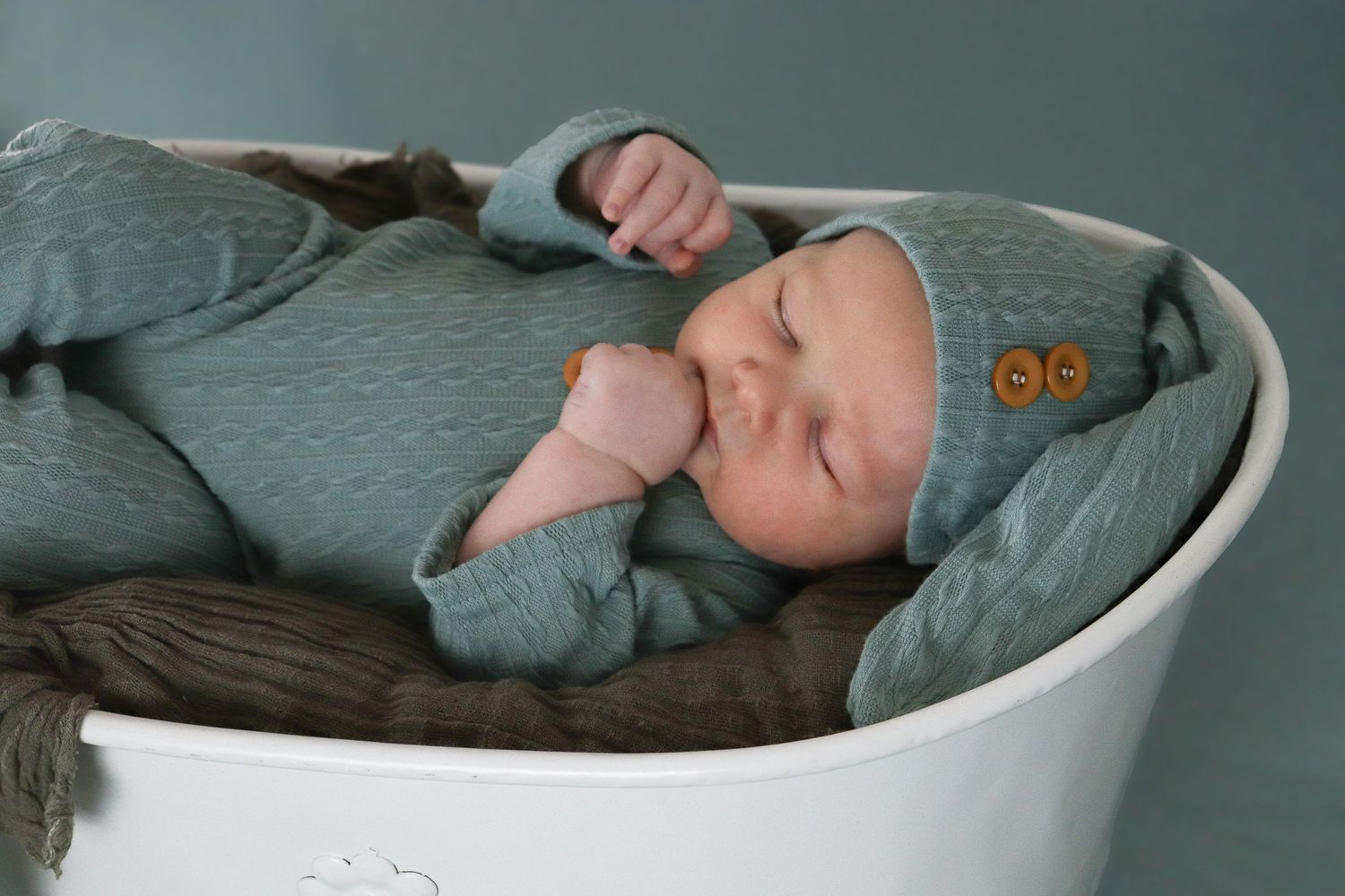 A sleeping baby wearing sage green clothing rests peacefully in a white modern baby bassinet.