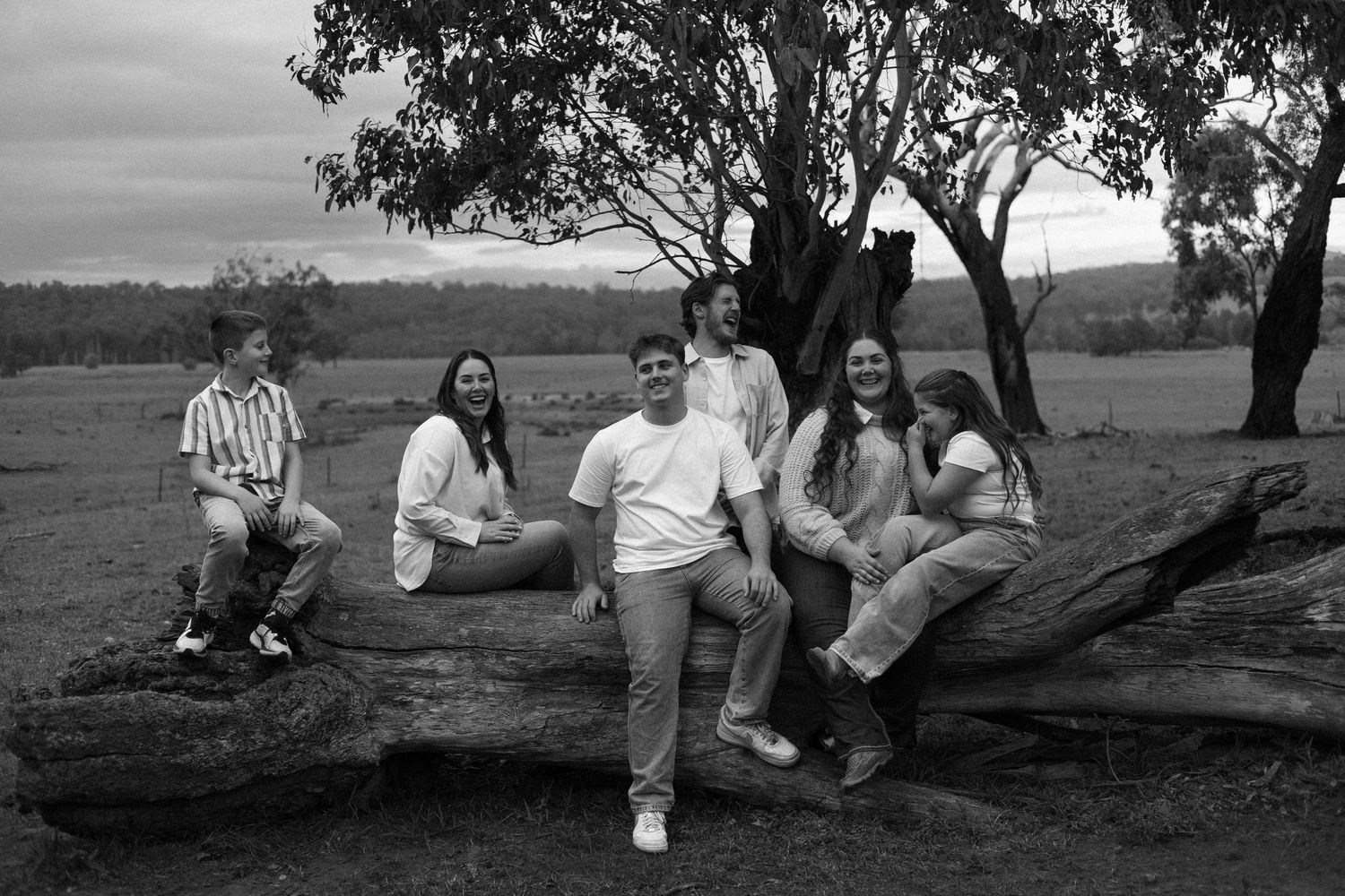 Group of five teenagers posing on a fallen log near a tree in a rural Australian landscape, black and white.