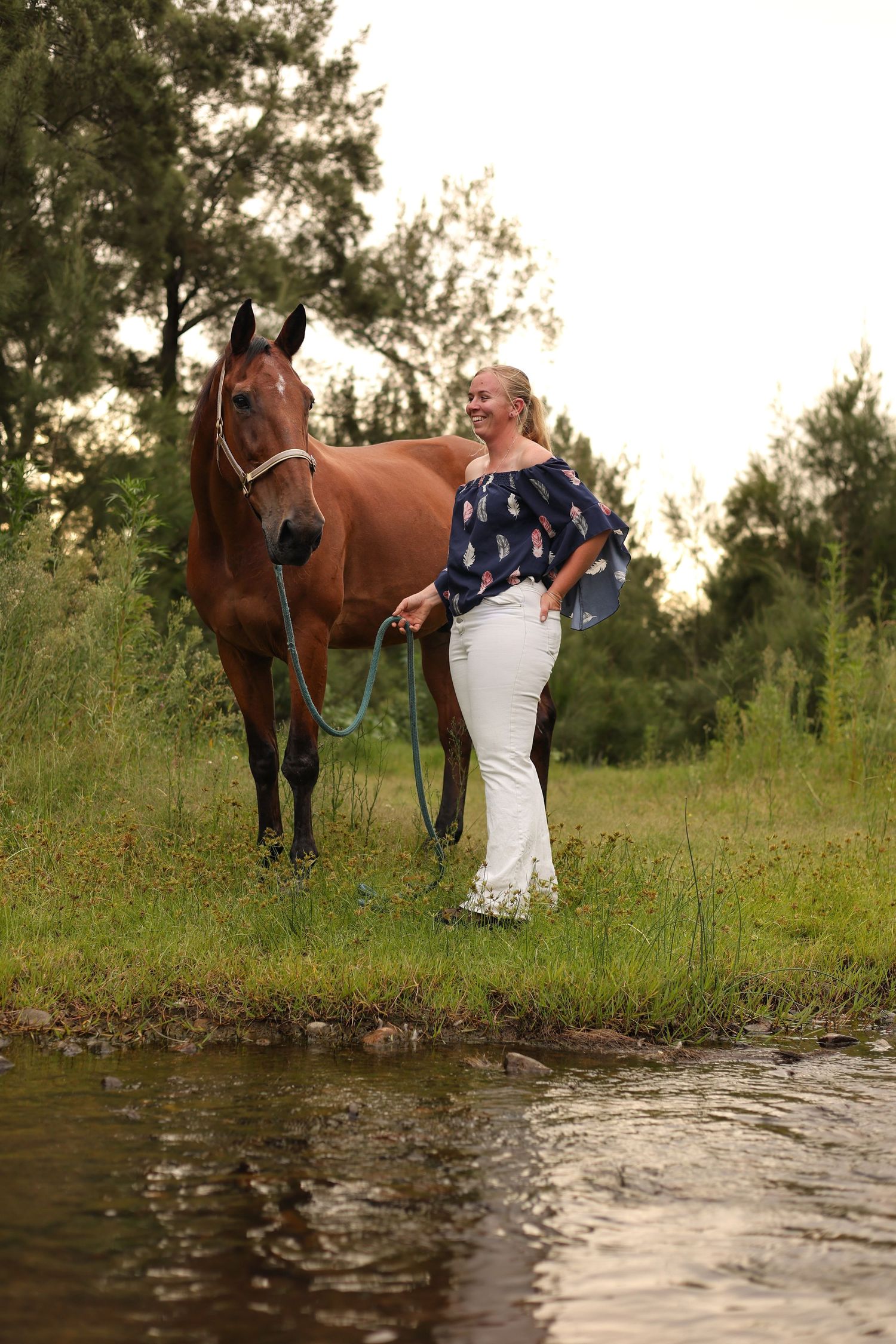 A couple poses with their brown horse by a stream in a scenic outdoor setting during golden hour.