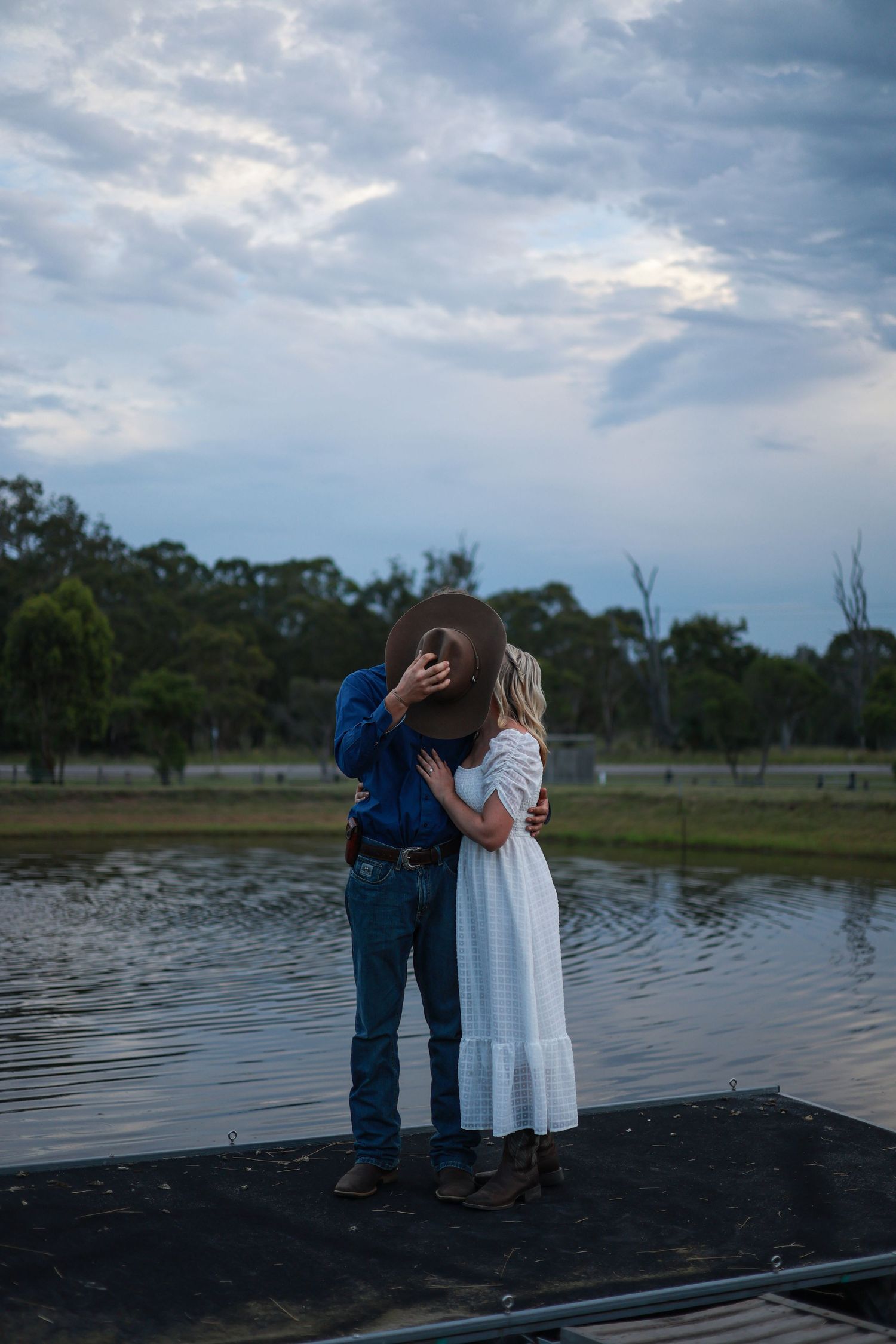 Two adults stand close together on a wooden dock overlooking a serene lake during an overcast evening.