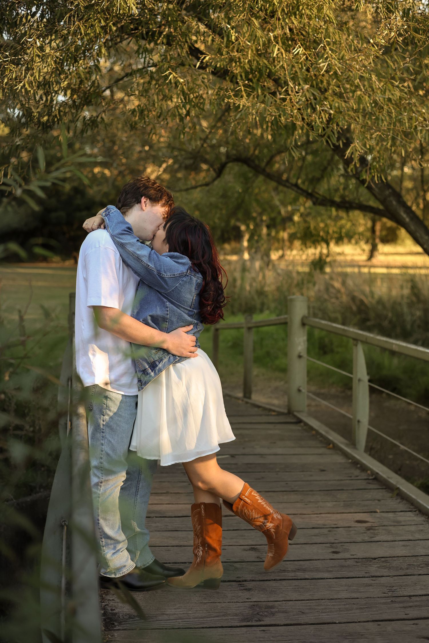 A couple sharing a romantic kiss on a wooden bridge under a willow tree, the woman wearing a white dress and brown cowboy boots.