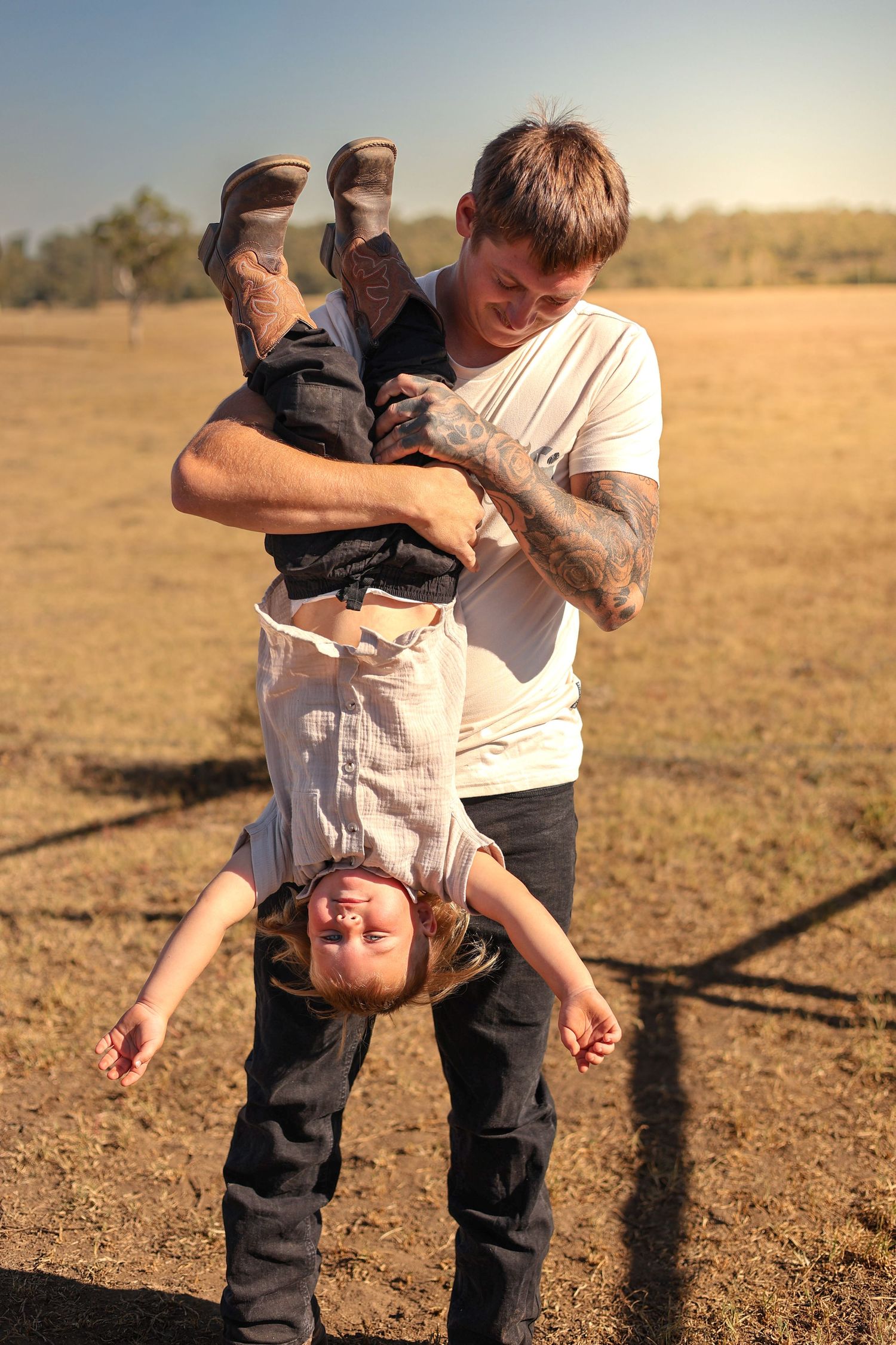 Tattooed father playfully holding young child upside down in sunny open field, both smiling and enjoying outdoor playtime.