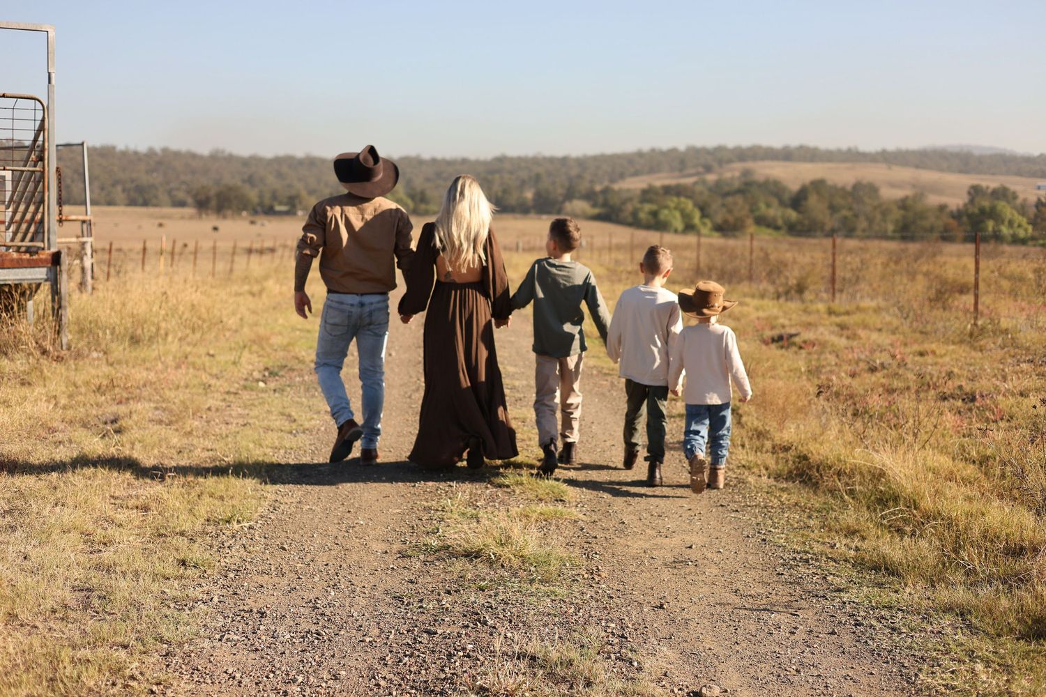 Family of five walking hand-in-hand down a rural dirt path surrounded by golden fields and trees.