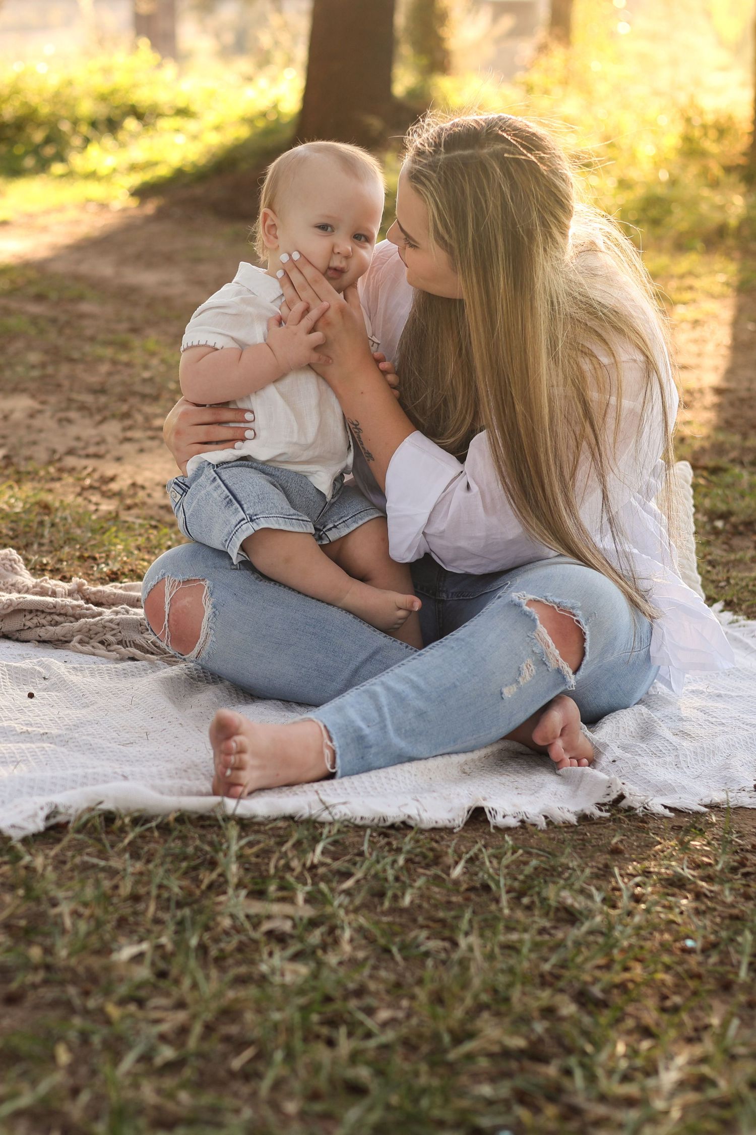 Young mother sitting barefoot on blanket in sunny park, kissing baby in jeans and white shirt.