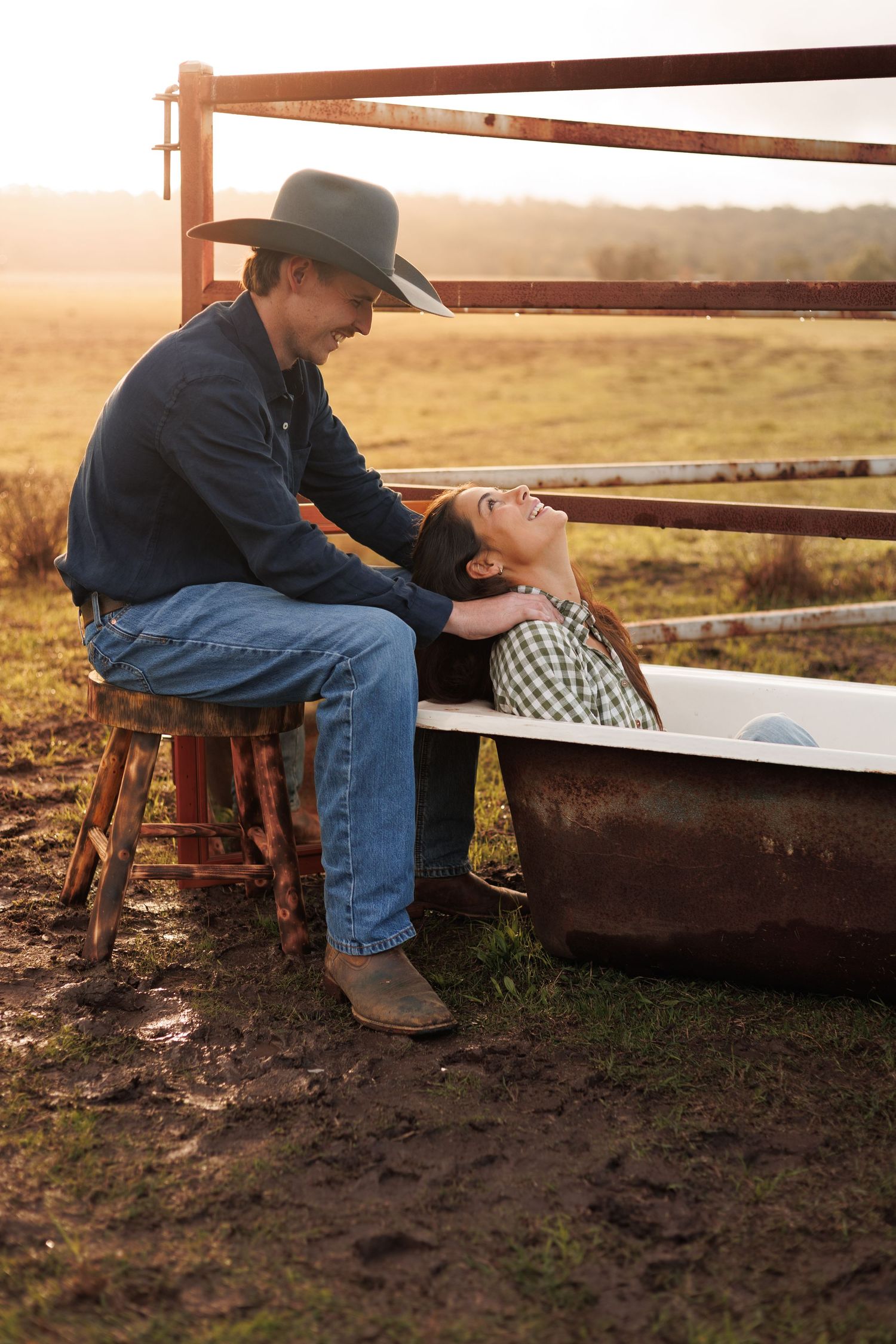 A couple shares a romantic moment at sunset, man in cowboy hat sitting by a rustic bathtub outdoors on a farm.