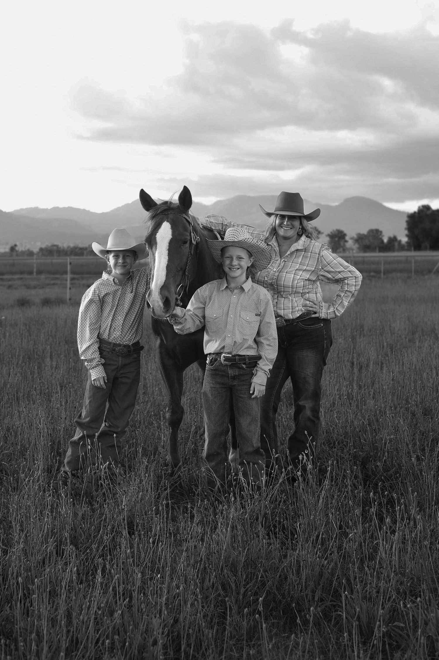 Black and white photo of two children and an adult in cowboy hats posing with a horse in a field.