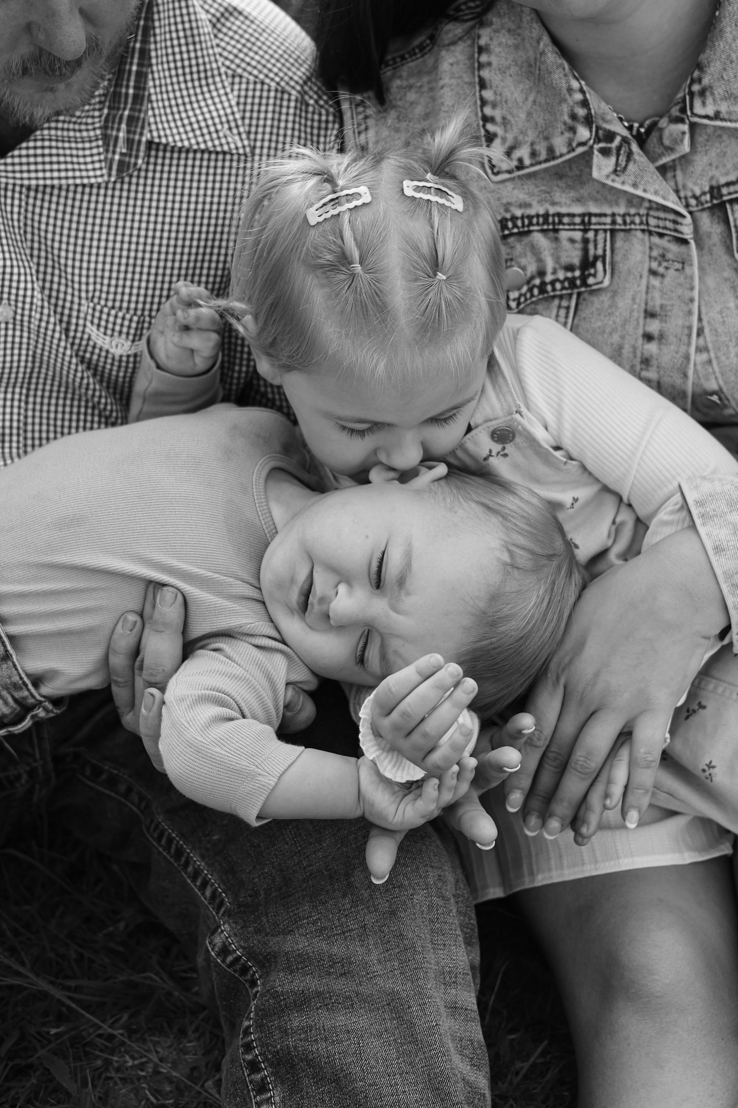 Black and white photo of two young siblings sharing a sweet kiss while sitting on parents laps outdoors.