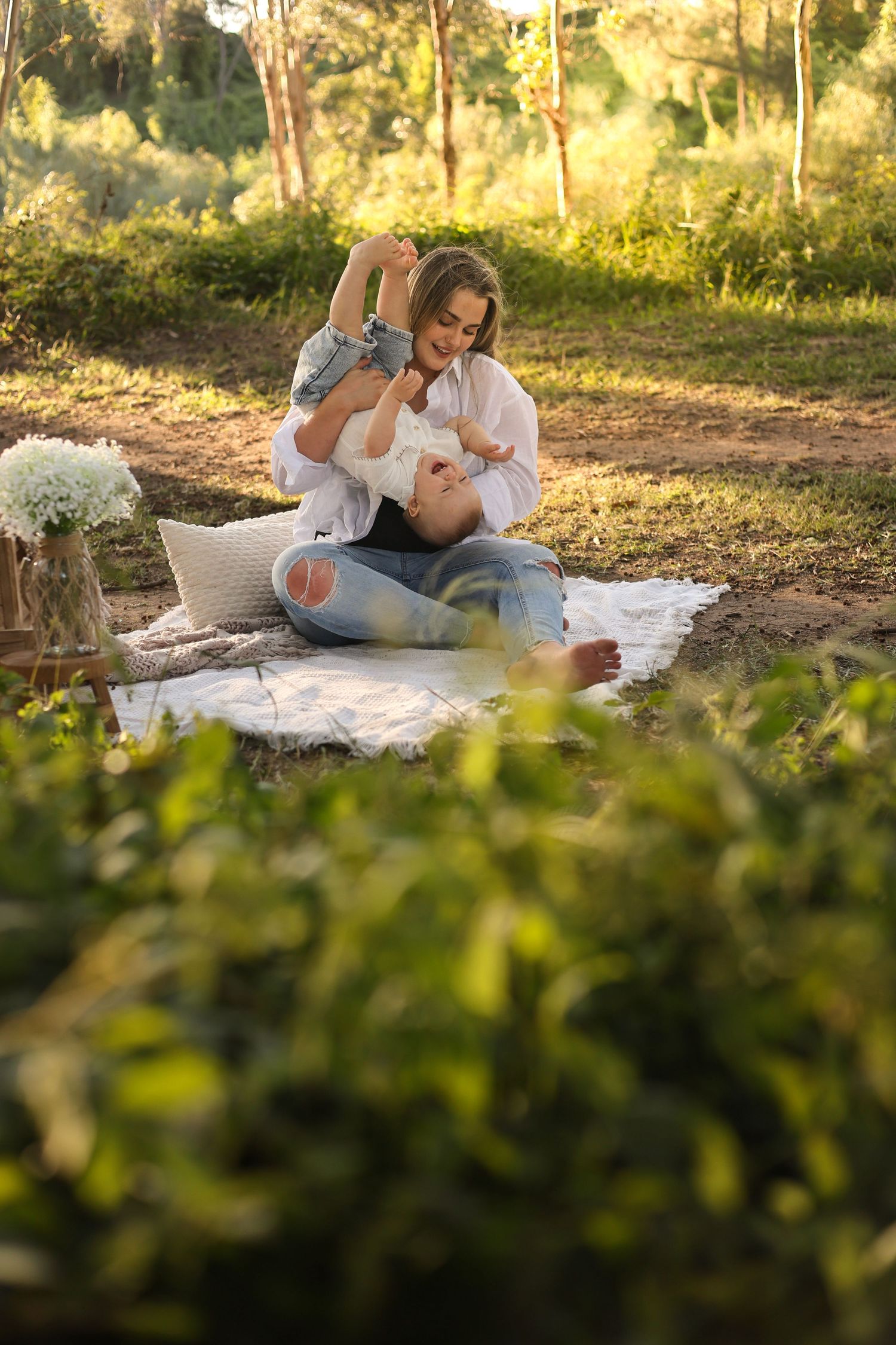 A mother sits cross-legged on a blanket outdoors, joyfully lifting her baby in a sunlit green park setting.