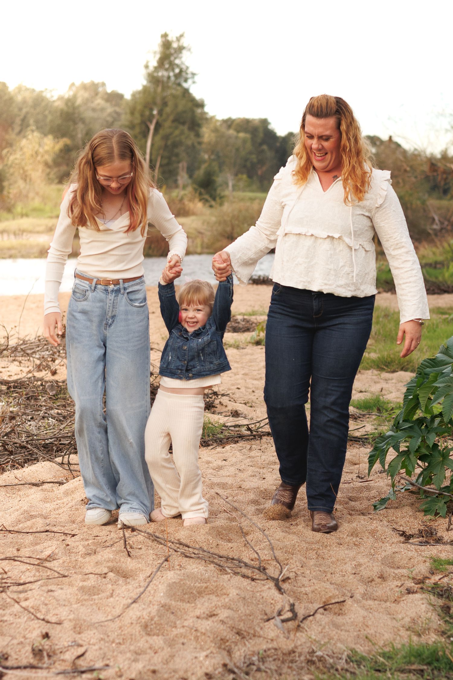 A mother and two children walk hand-in-hand on a sandy riverbank surrounded by nature.