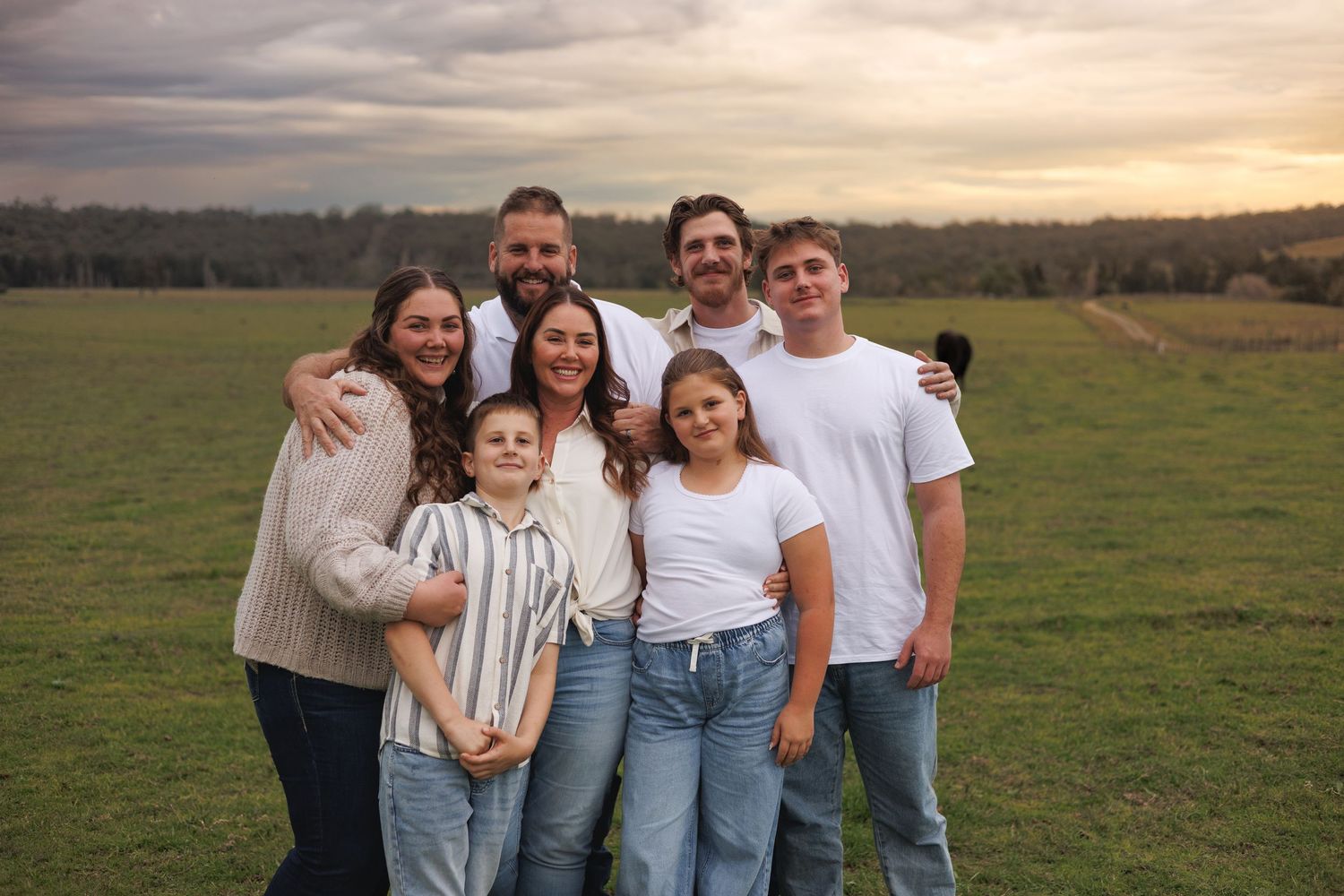 Family of seven smiling together in a green field at sunset, wearing casual jeans and tops.