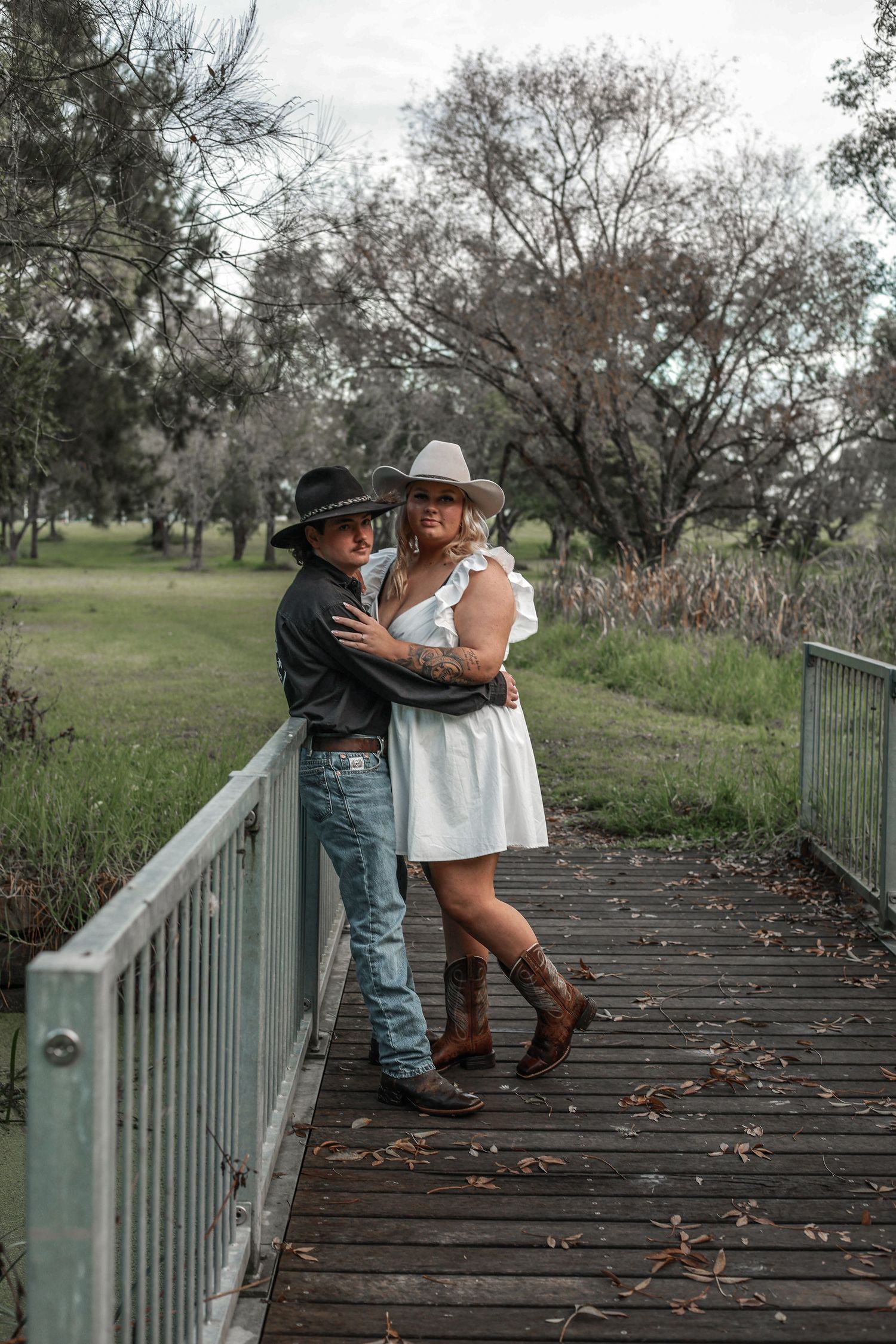 A romantic couple in western attire embraces on a wooden bridge surrounded by trees on an autumn day.