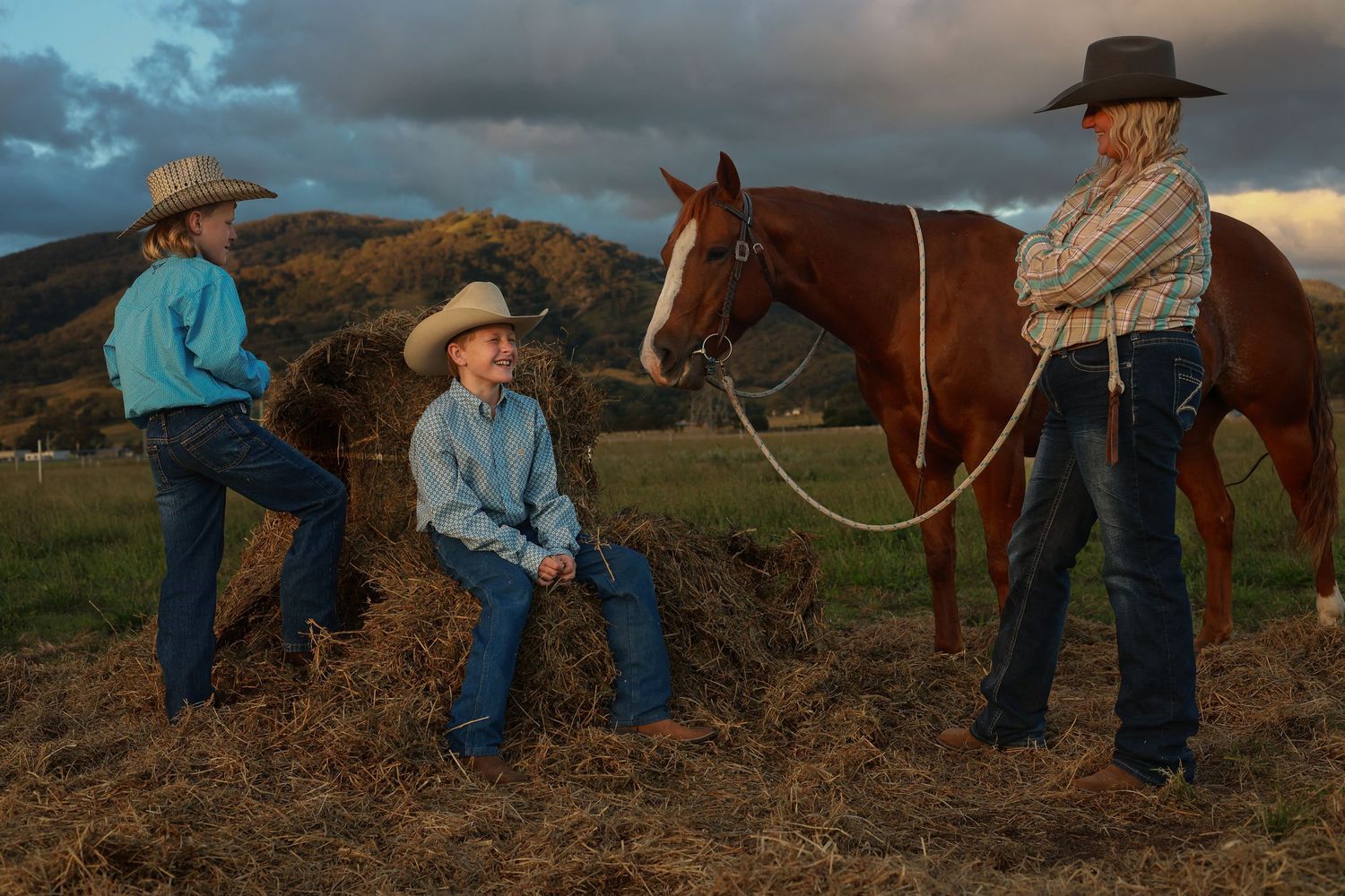 Two children and an adult in cowboy hats stand near a chestnut horse on a ranch at dusk.