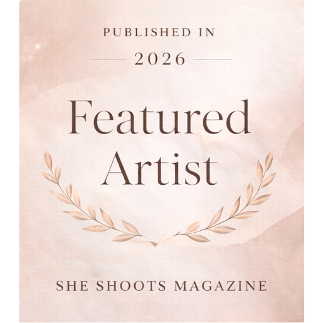 She Shoots Magazine Featured Artist badge published in 2026 with elegant floral laurel wreath design on soft pink background.