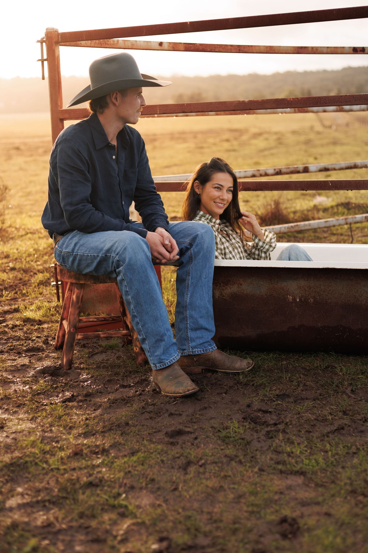 A cowboy in a hat and woman in jeans relax by a rustic trough at golden hour on a farm.