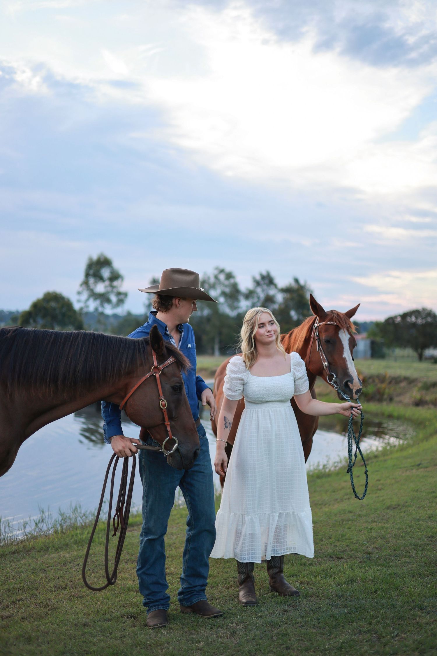 Couple and horse stand together by a scenic lake at sunset during a romantic countryside photo session.