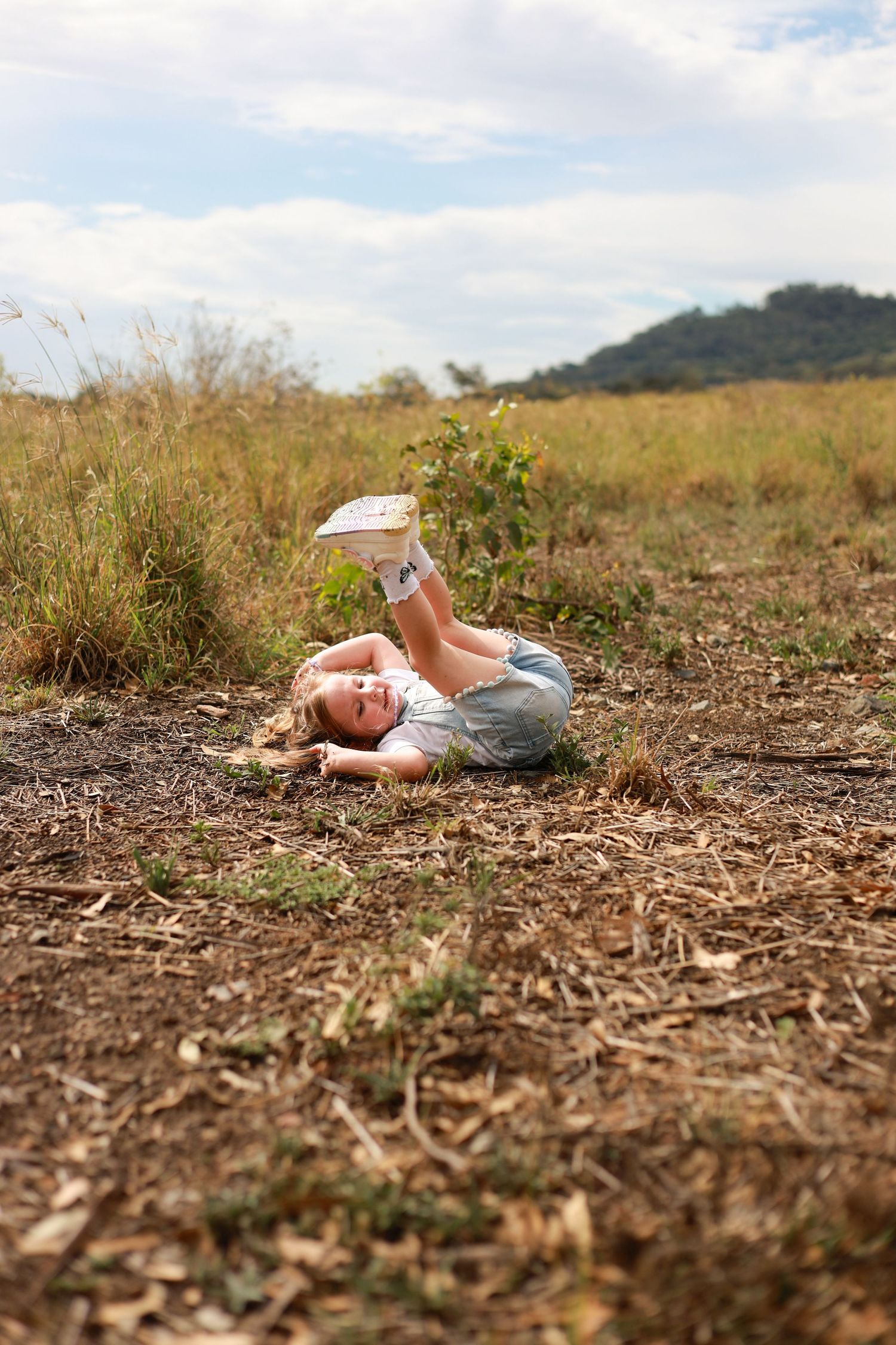 Young girl lying on dry grass in a field, reading a book outdoors on a cloudy day.