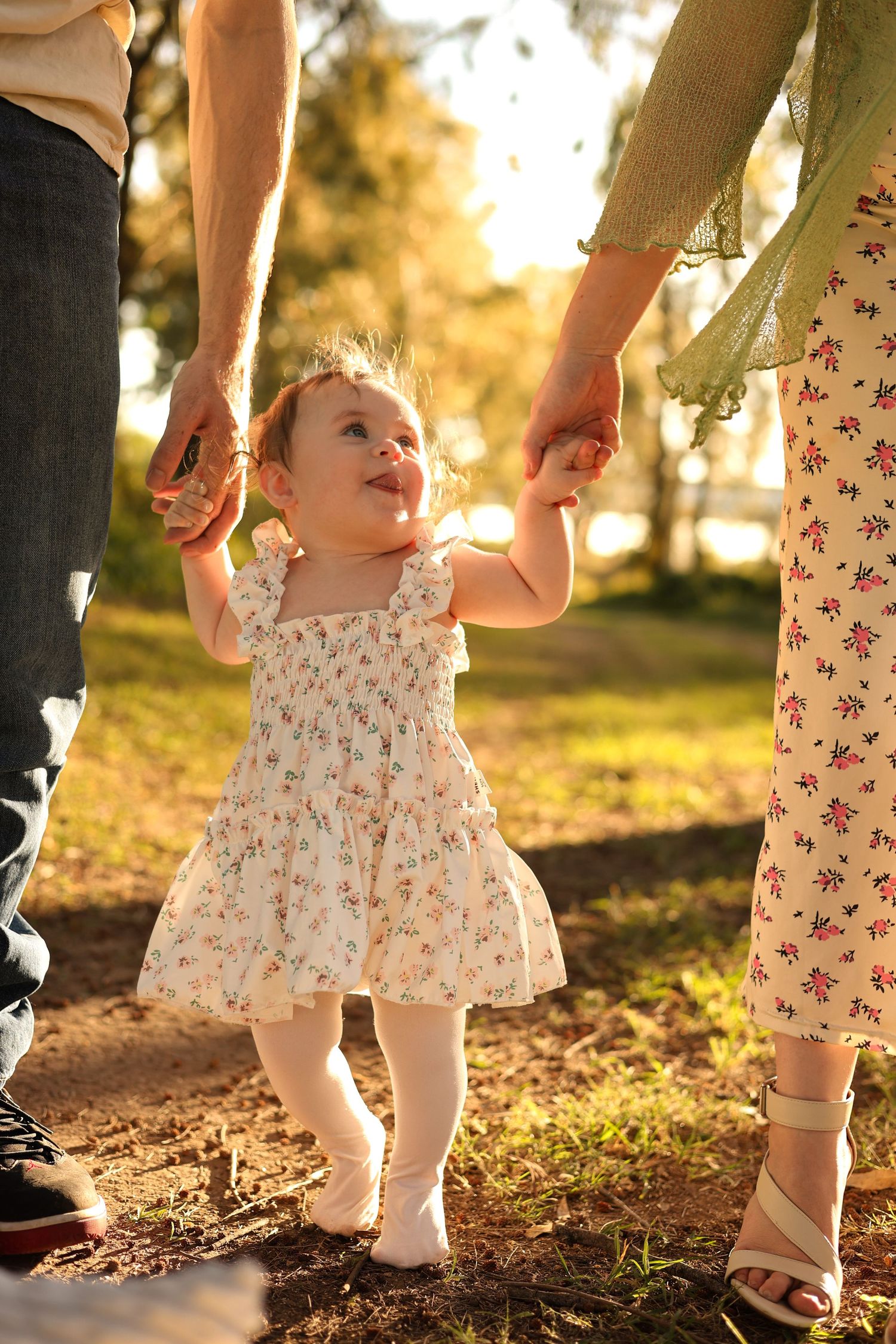 A toddler girl in a floral dress holds her parents hands while learning to walk in a sunlit park.
