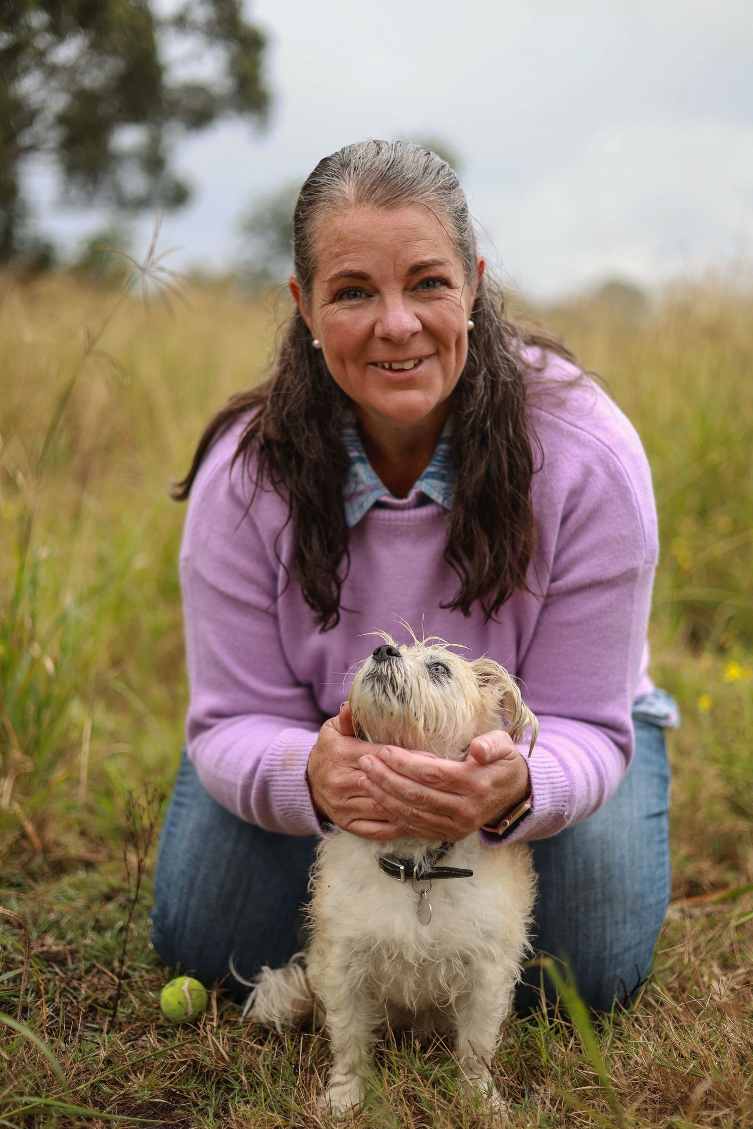 A person in a pink sweater plays with a small white fluffy dog in a grassy field on a rainy day.
