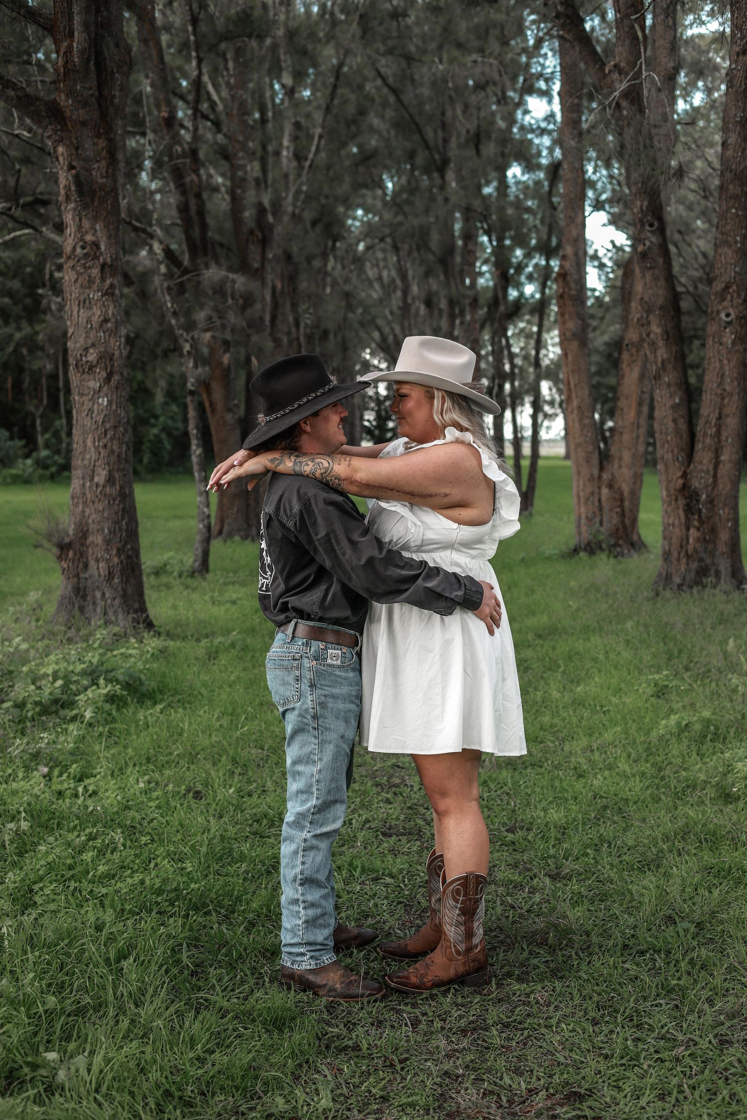 A couple in cowboy boots and casual attire share an intimate embrace among tall pine trees in a grassy forest setting.