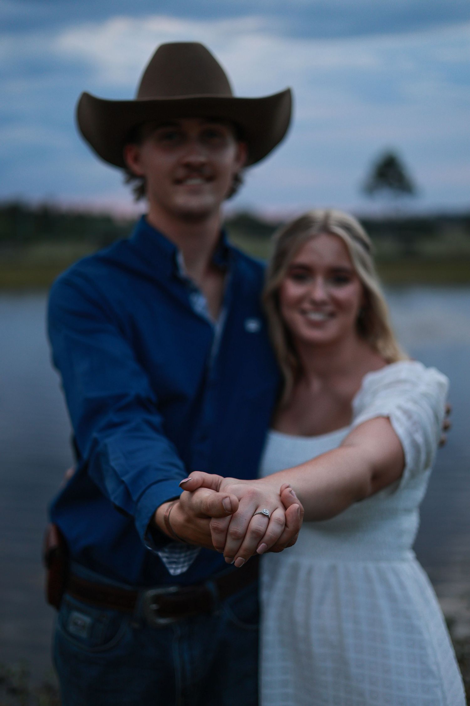 A couple in western attire hold hands and pose together by a scenic lake at sunset.