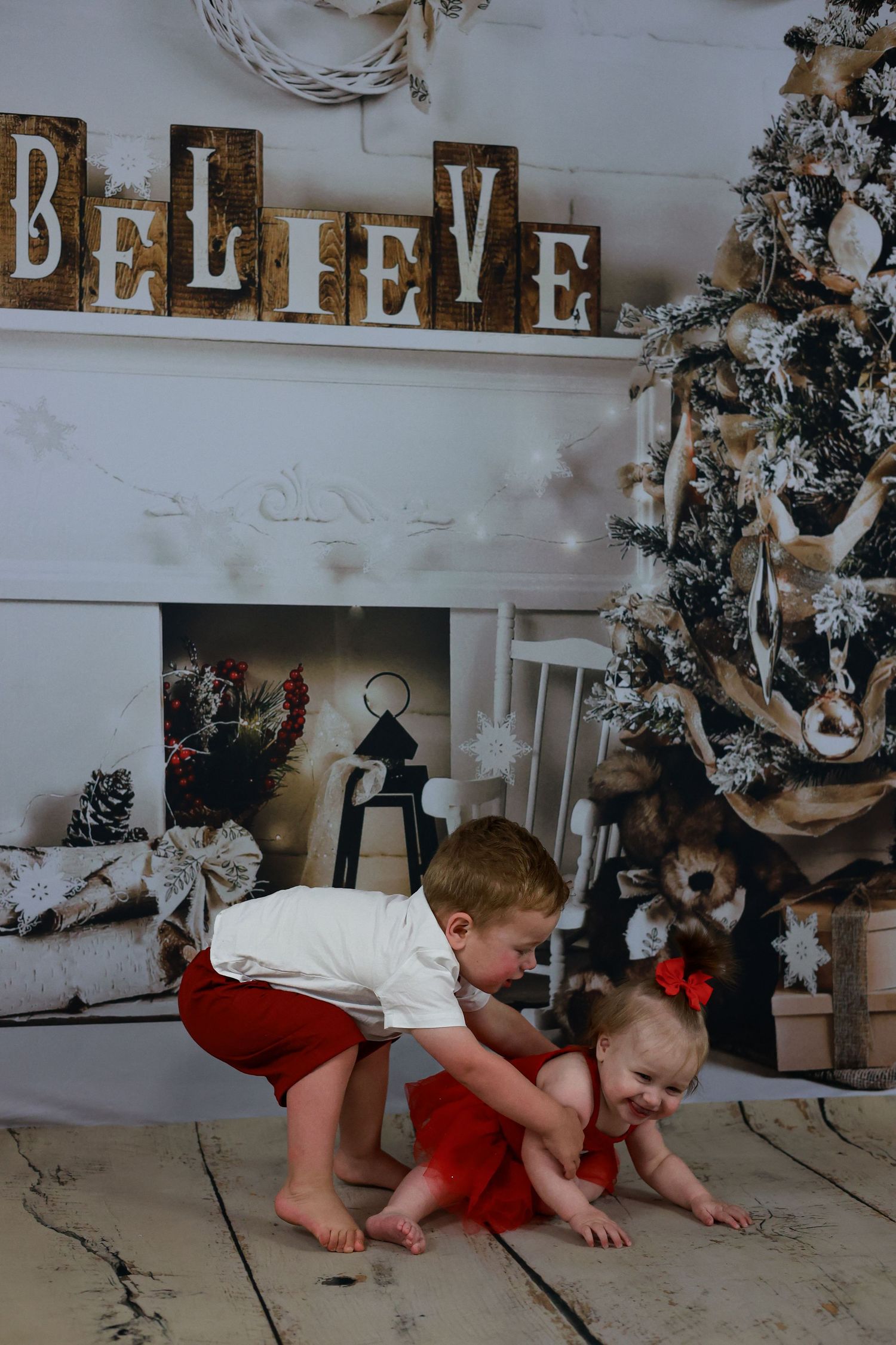Two children play together in front of a Christmas tree and fireplace decorated with rustic 'BELIEVE' letters.
