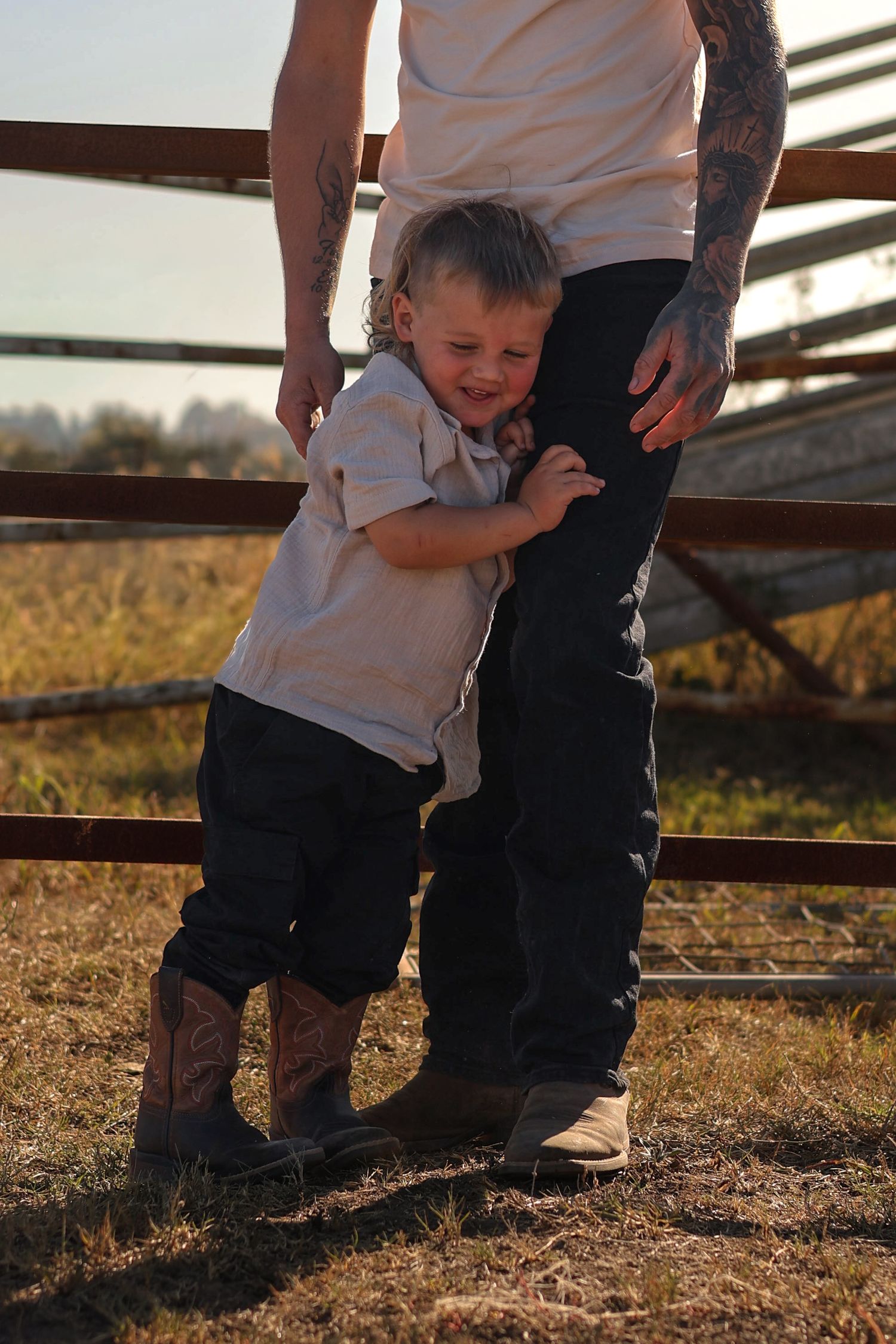 Young boy in cowboy boots hugging an adult's legs near a rustic fence in a sunny field.