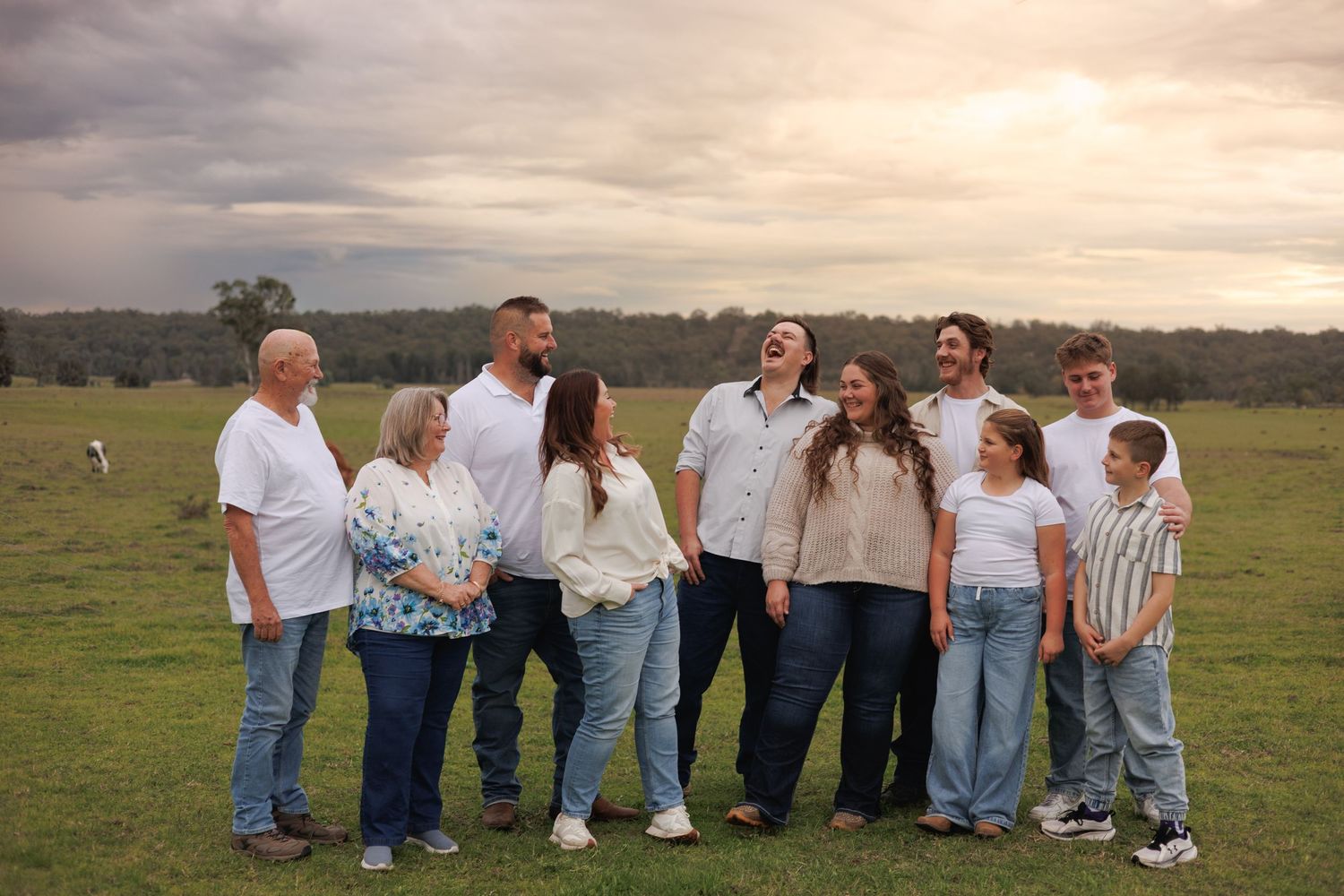 A joyful family of nine poses and laughs together in a green field under a cloudy sky, wearing white tops and jeans.