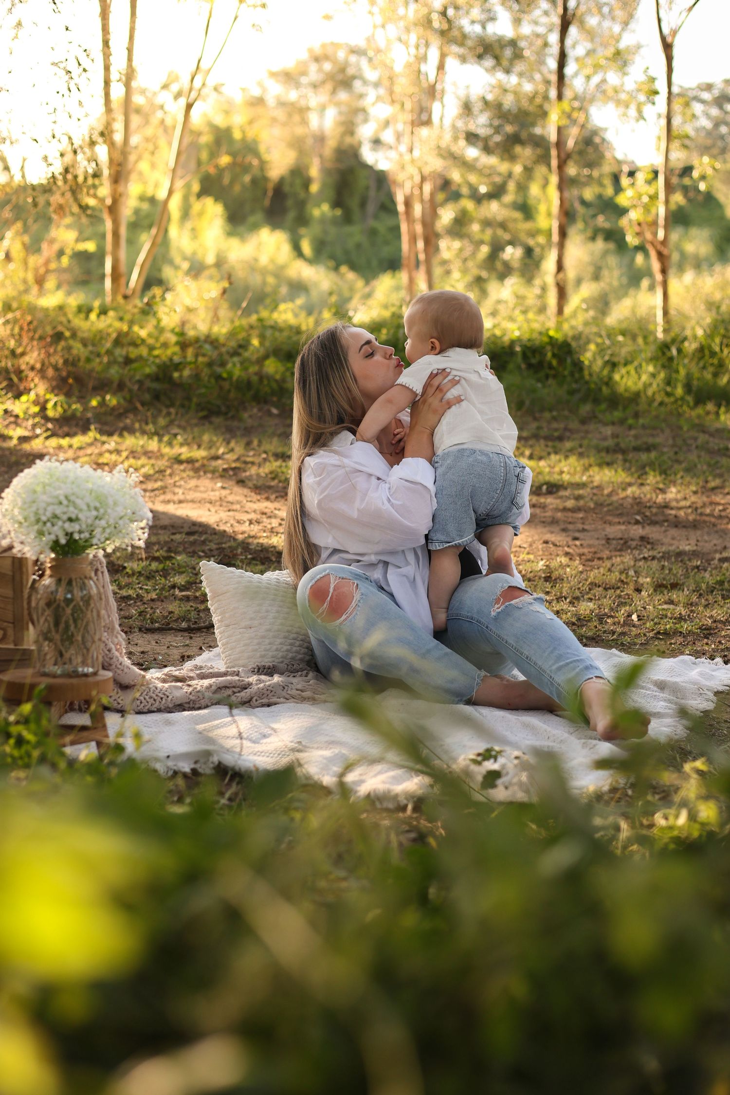 A mother kisses her baby on a picnic blanket outdoors, surrounded by lush greenery in warm golden sunlight.