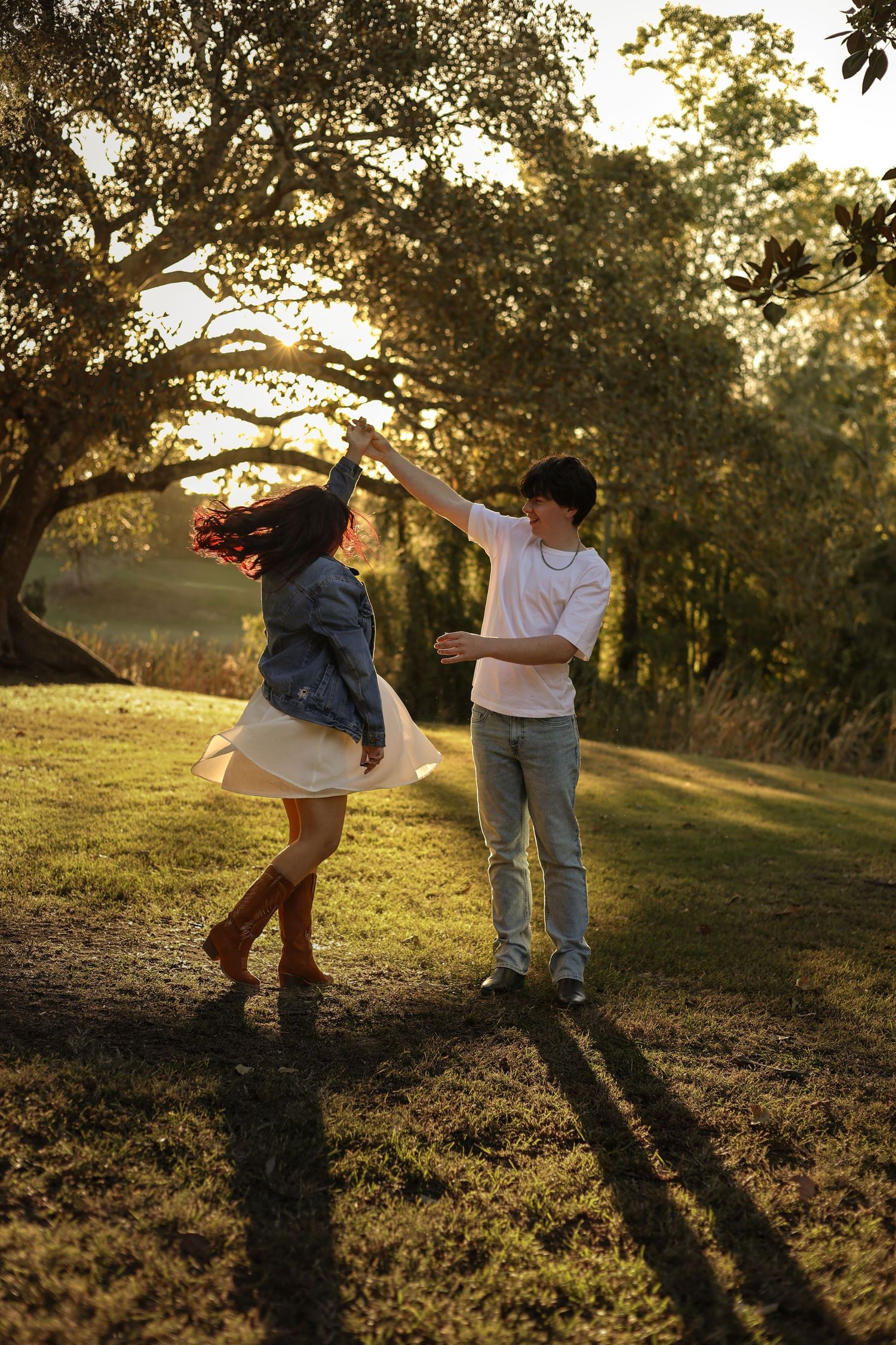Couple dancing outdoors at golden hour, woman in denim jacket spinning in white skirt, man in white shirt, under large oak trees.