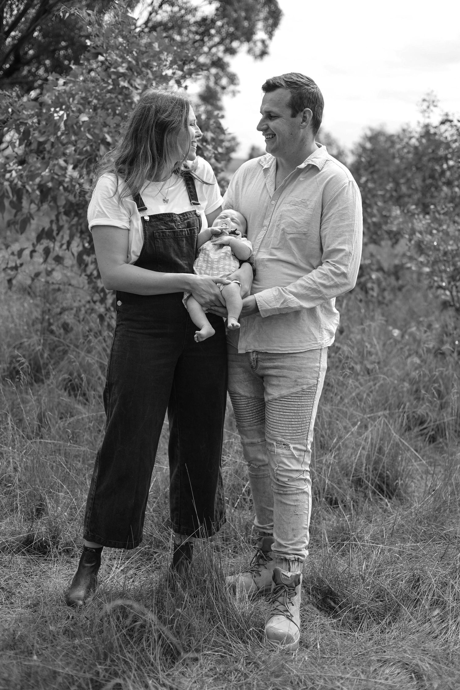 Black and white photo of a couple smiling at each other while holding their newborn baby outdoors.