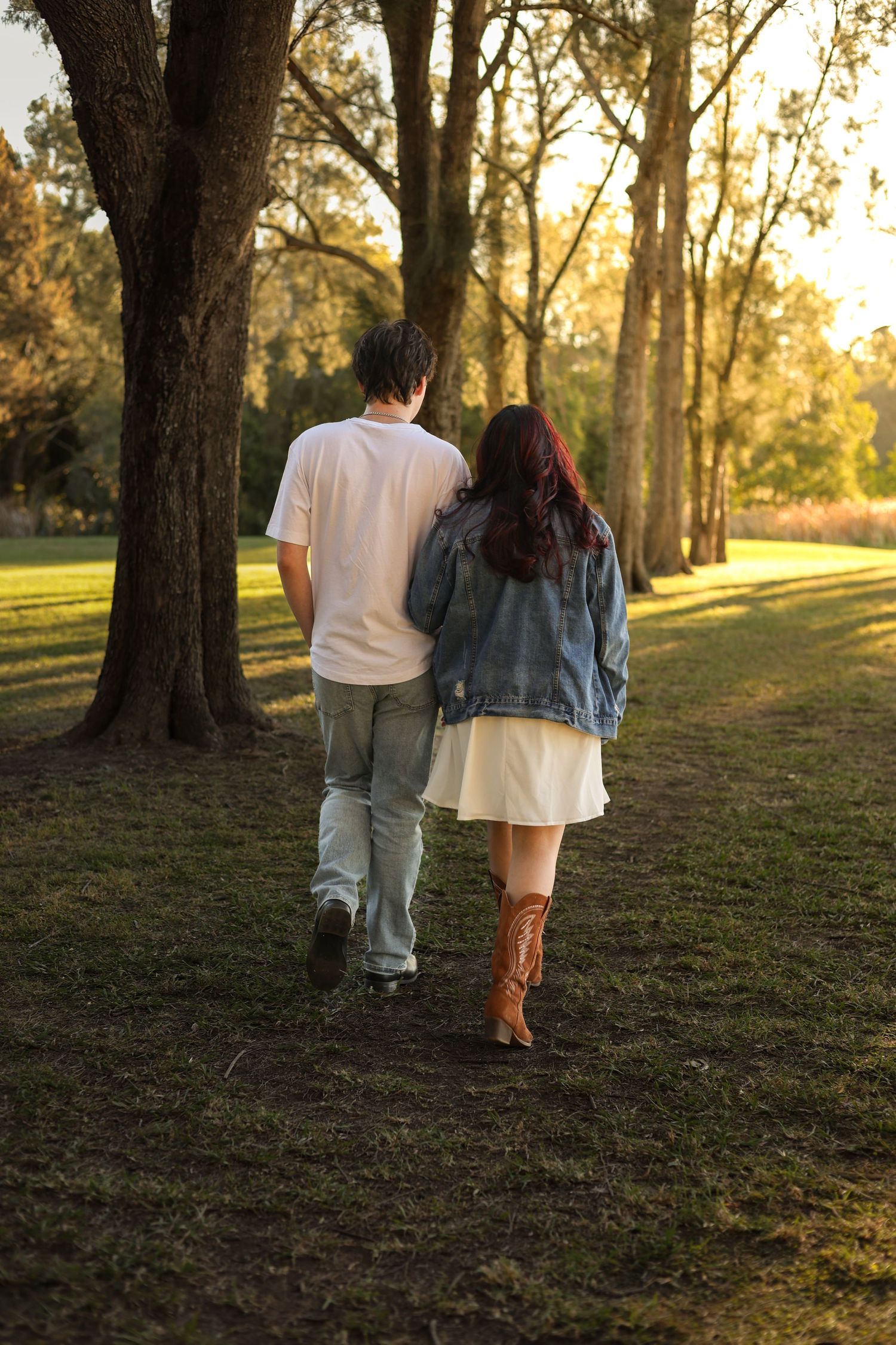 Couple walking hand in hand through a sunlit park at golden hour, woman in denim jacket and cowboy boots.