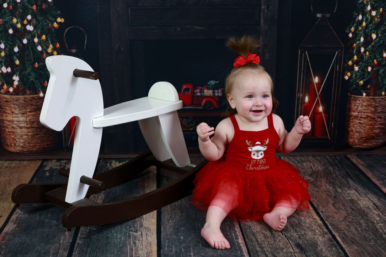 Smiling baby in red holiday dress sitting next to white wooden rocking horse with Christmas decorations in background.