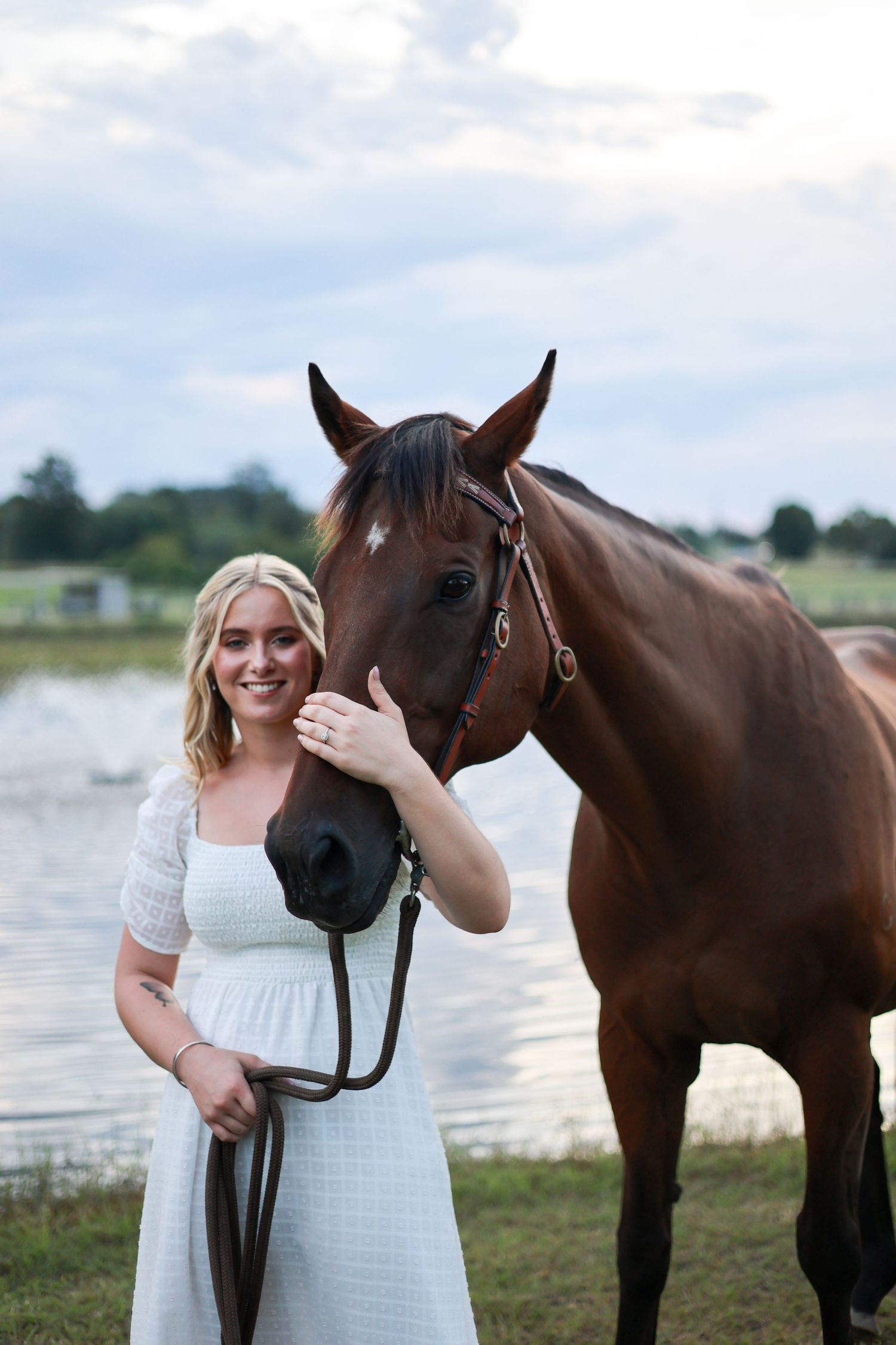 A beautiful brown horse standing next to a pond with its handler in a white dress on a sunny afternoon.