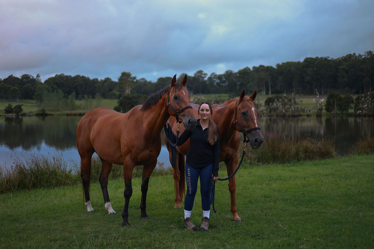 Two  horses stand with someone on grassy field next to a pond at dusk.