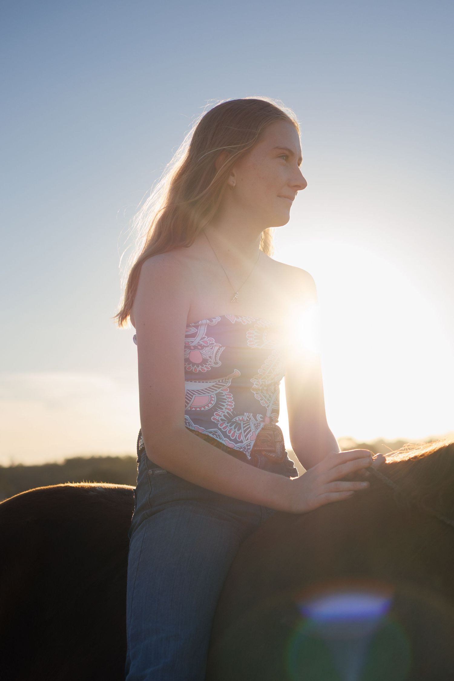 A young woman with long hair rides a horse bareback at sunset, with golden sunlight glowing behind her.